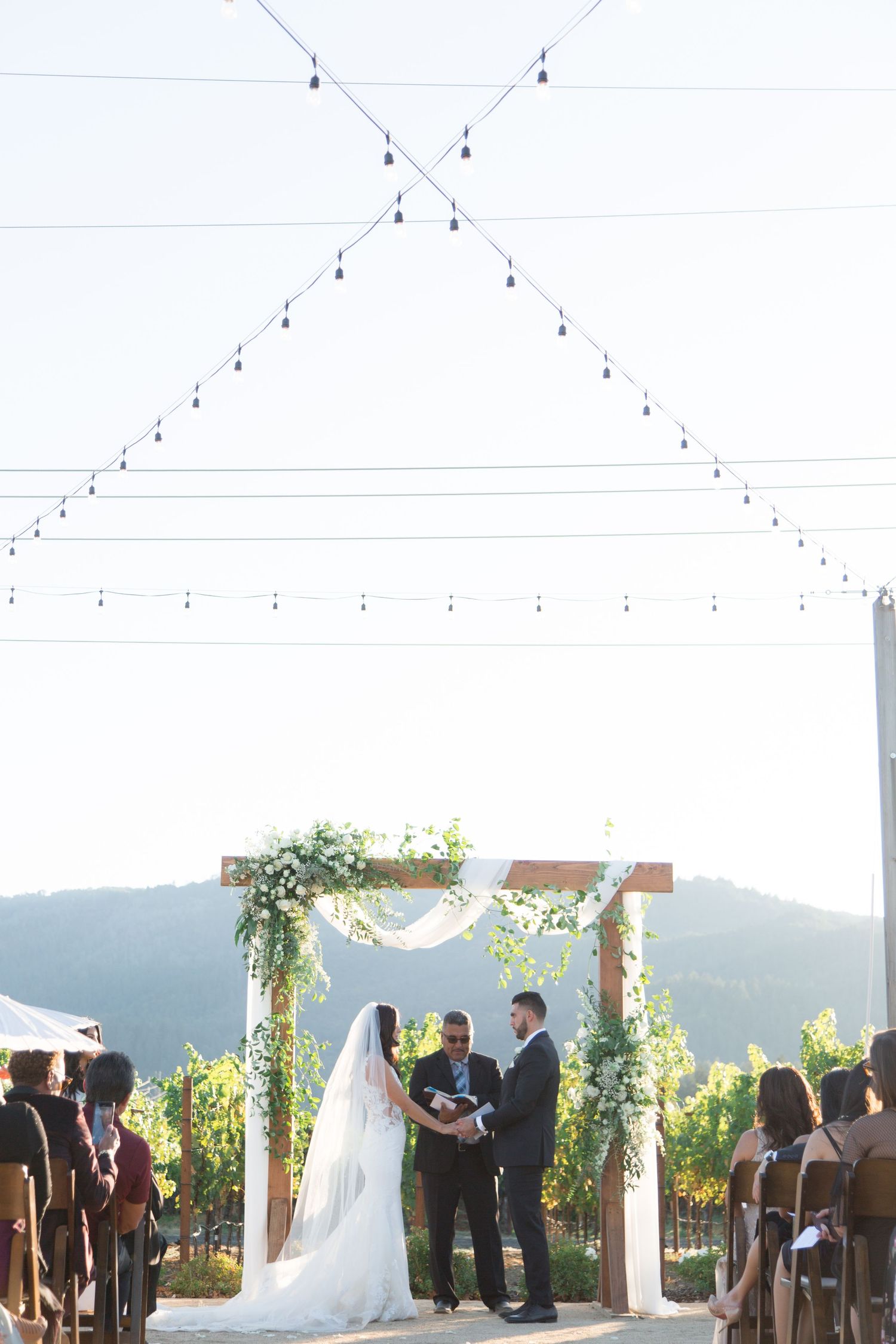 Wedding ceremony under a floral arch with mountain views and string lights overhead.