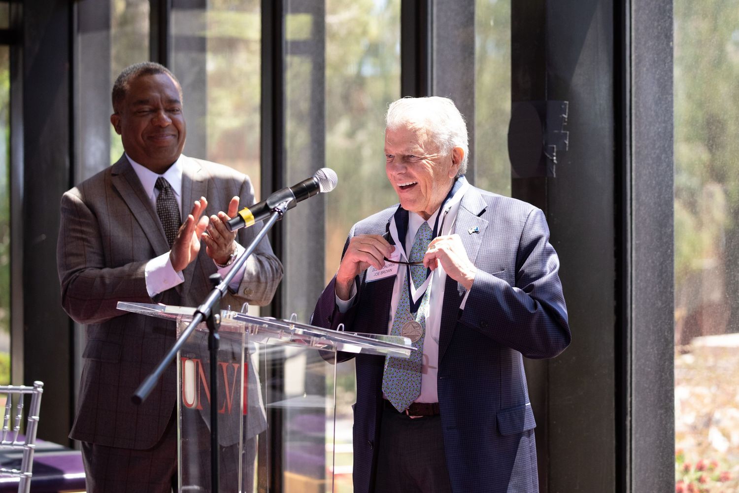 Two speakers share microphone at podium during presentation in front of windows.