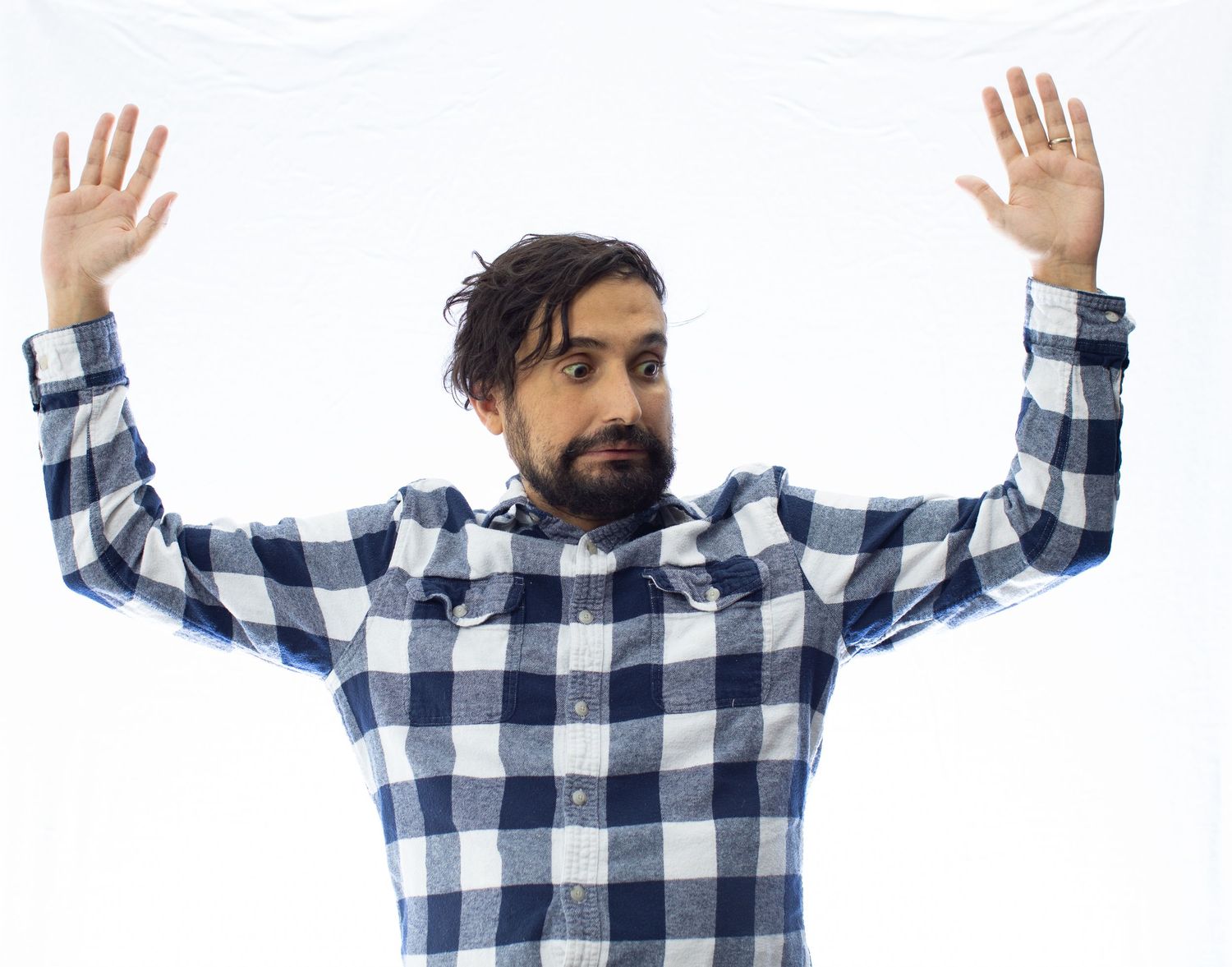 Person in blue plaid shirt raising hands in surrender pose against white backdrop.
