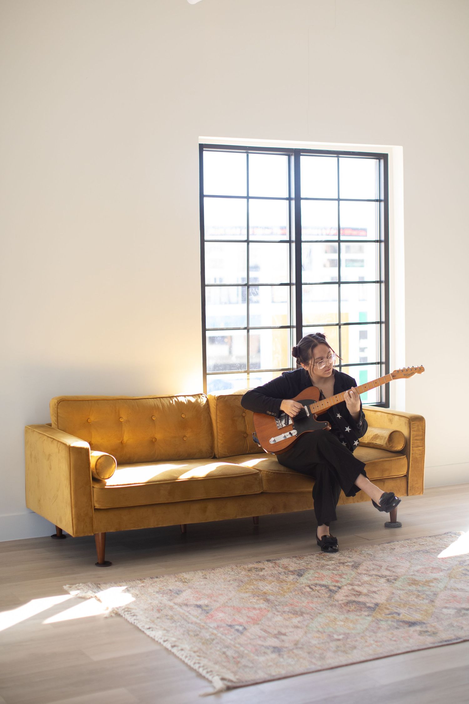 Musician plays guitar while sitting on yellow velvet couch near bright window.