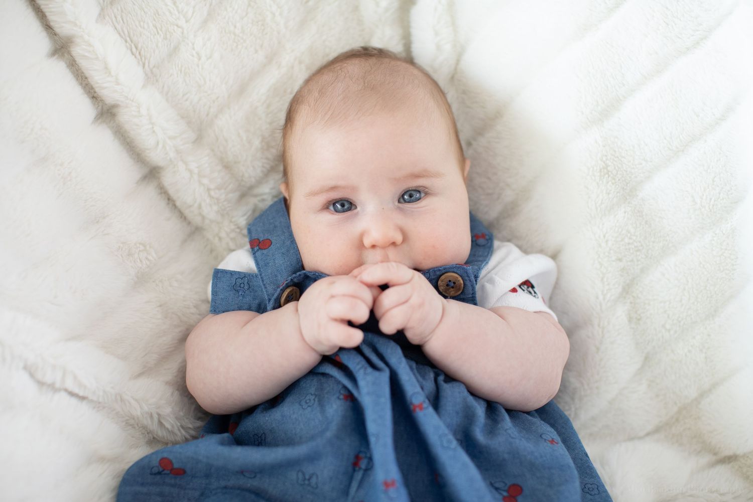 A baby wearing a blue dress lies on a white quilted blanket while chewing on their fingers.