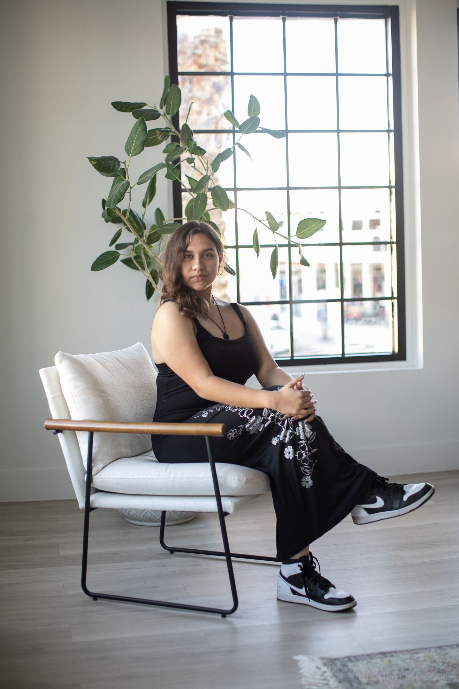 Person in black outfit poses on modern chair by window with large indoor plant.