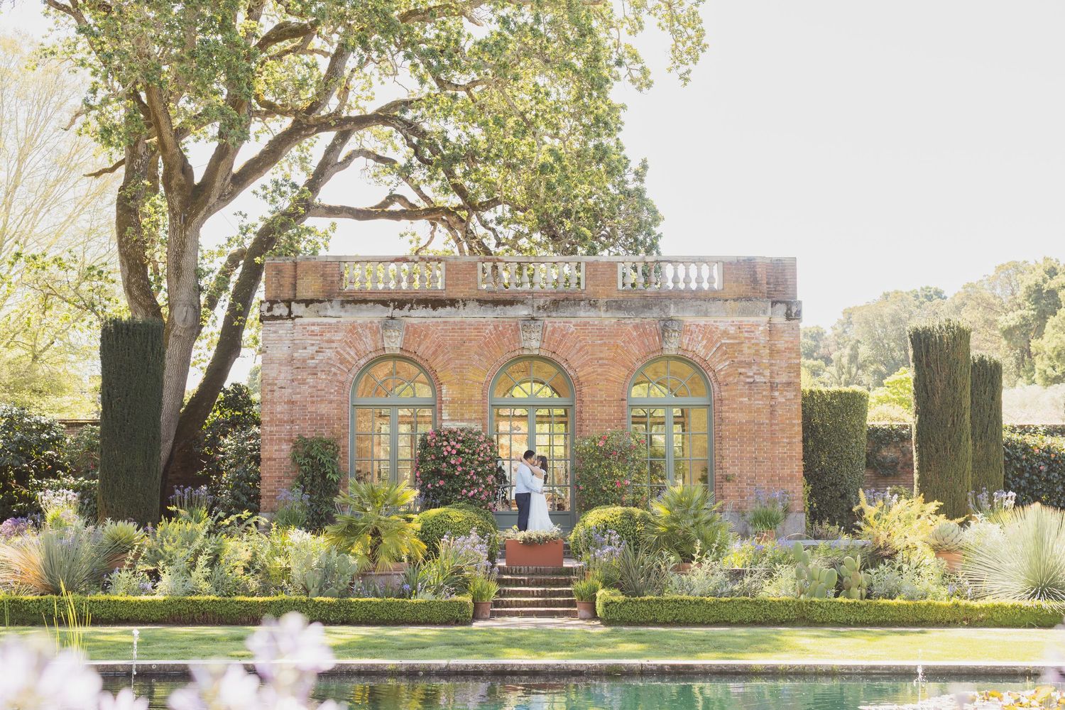 Couple embracing at elegant brick orangery wedding venue surrounded by formal gardens and reflecting pool.
