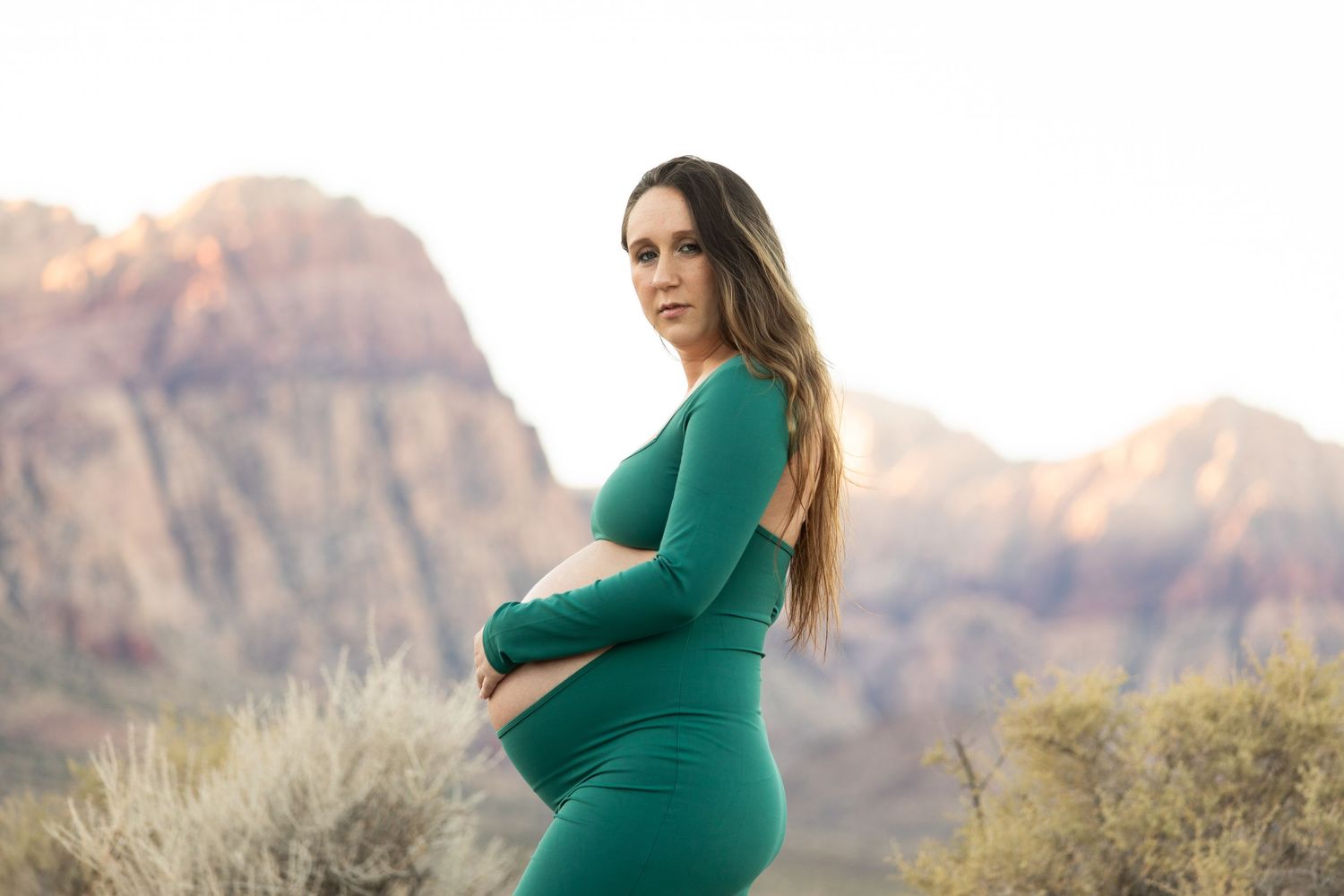 Expectant mother in green dress poses against red rock mountains during sunset.