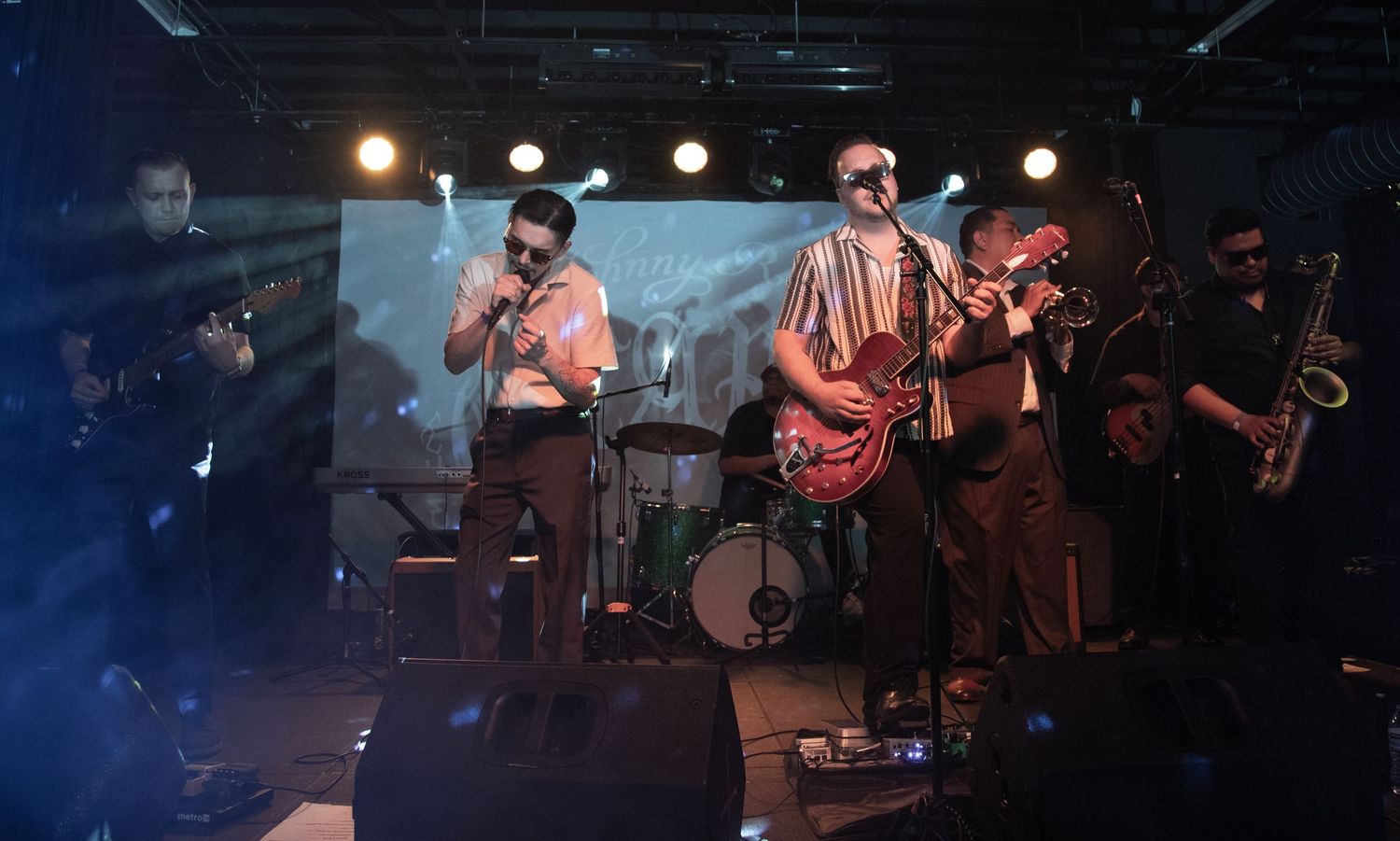 Full band performs on stage with instruments under theatrical lighting setup.