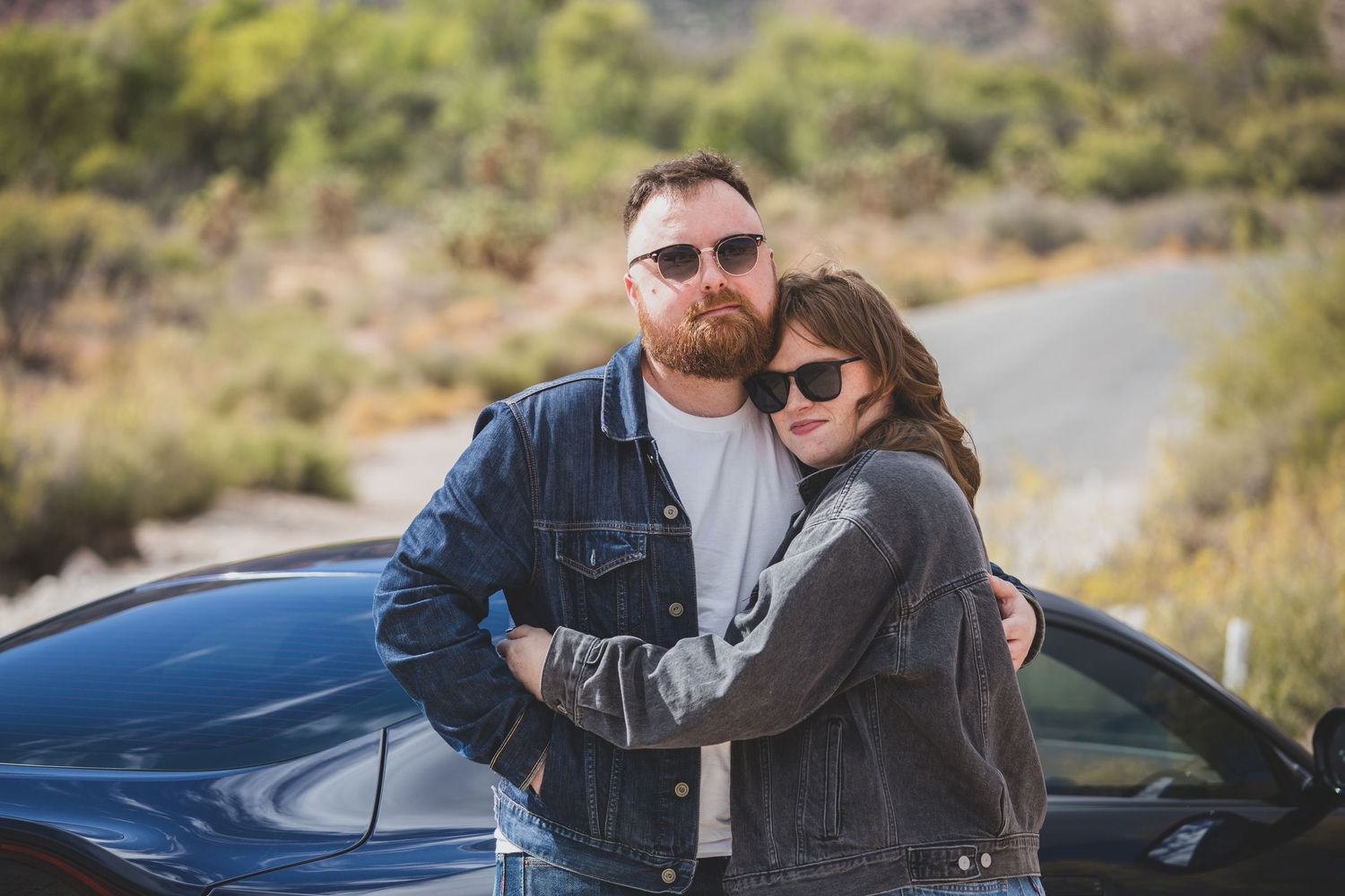A couple embracing next to a blue car on a desert road.