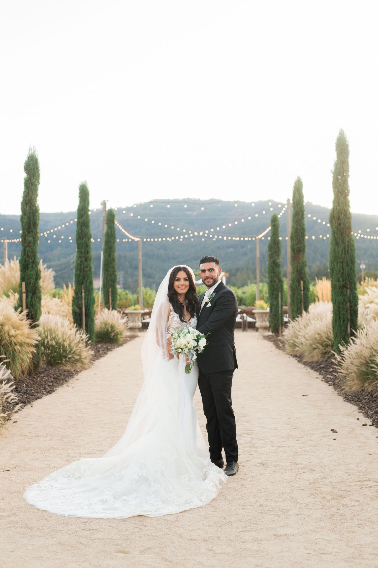 Mediterranean-style wedding venue features tall cypress trees and string lights along a path.
