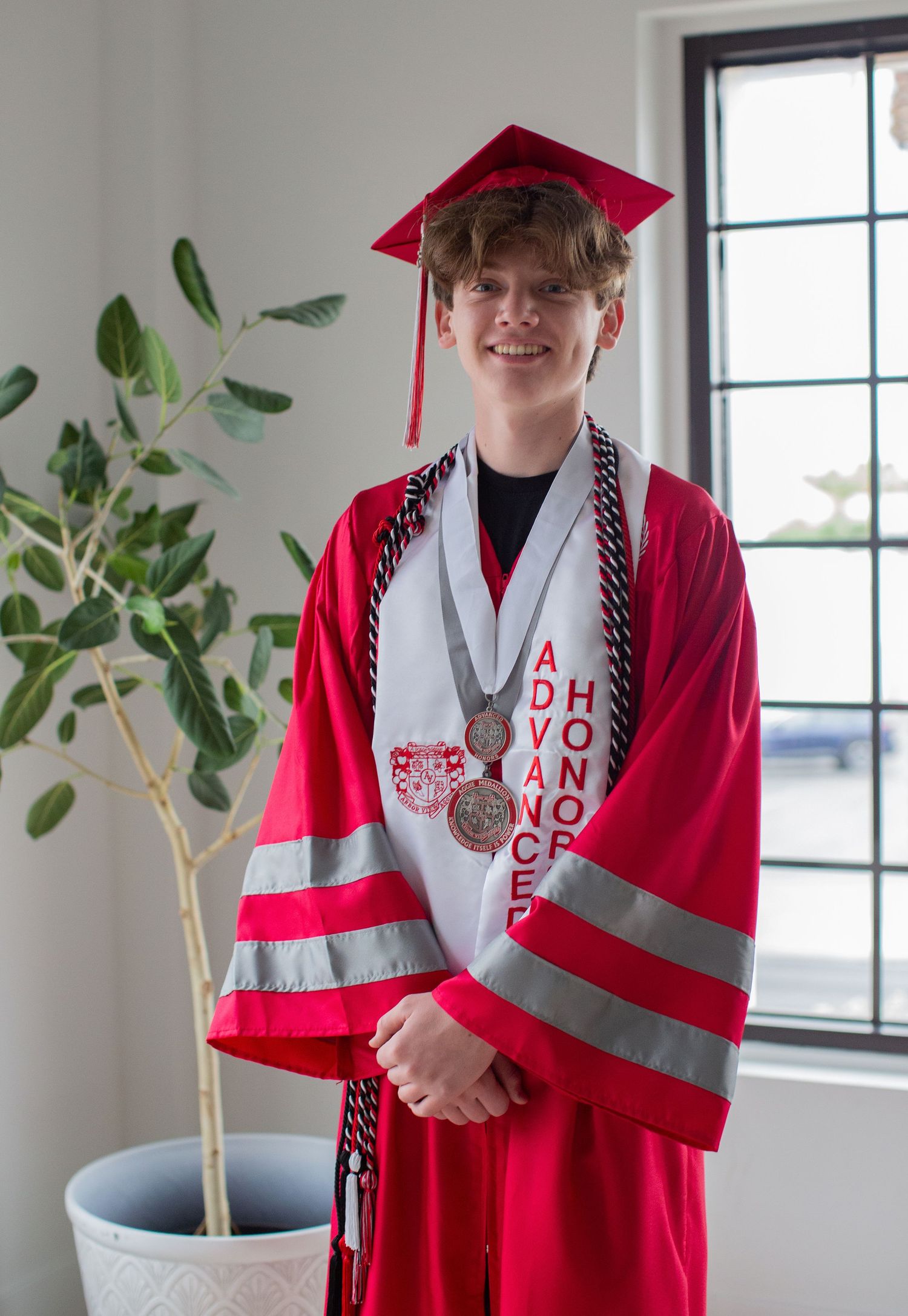 Graduate in red and white academic regalia poses by window with decorative plant.