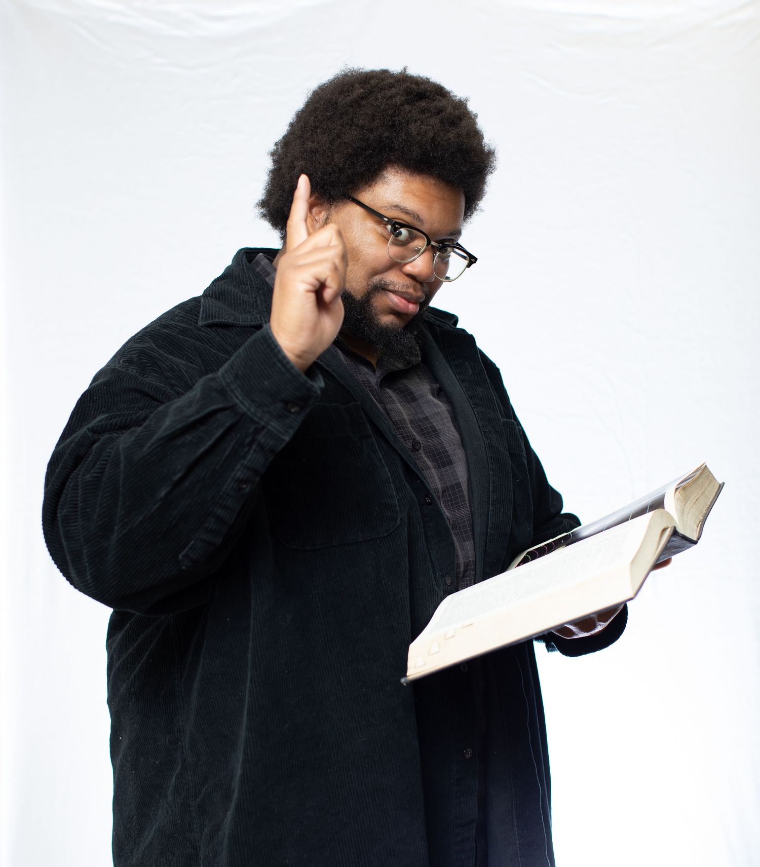 Person in black coat holding book and raising finger in thoughtful gesture against white background.