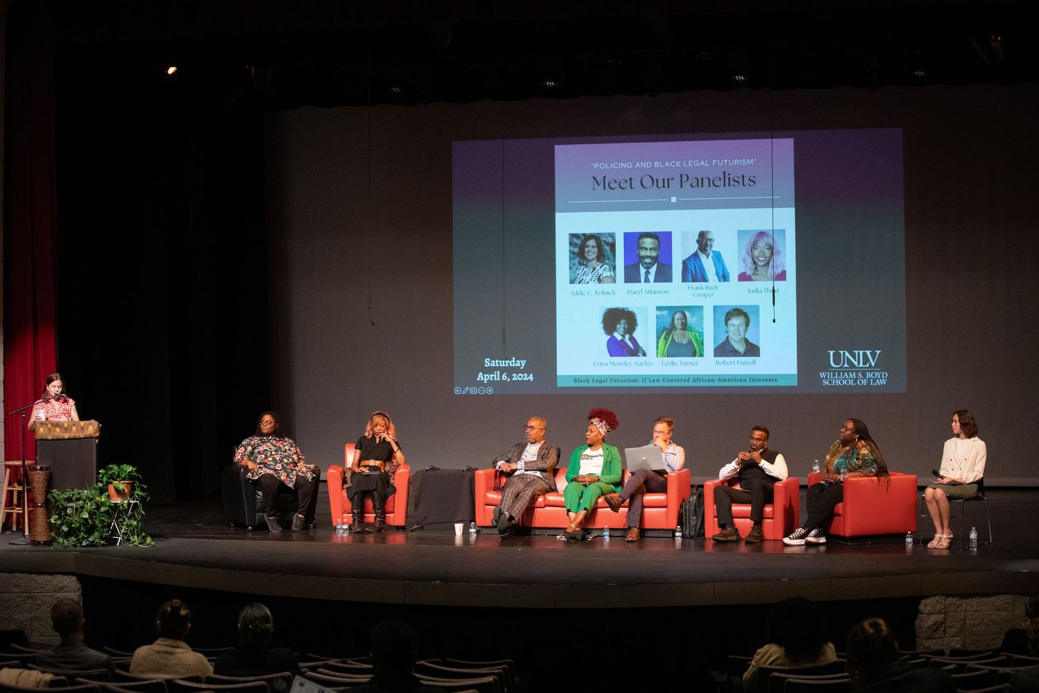 Panel discussion featuring multiple speakers seated on stage with red chairs.
