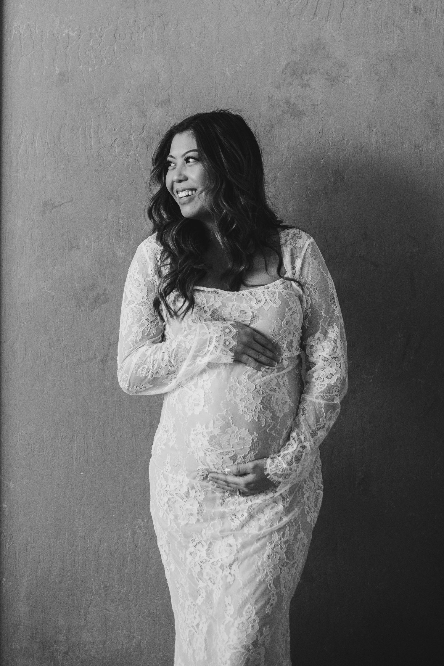 Expectant mother in white lace dress poses against textured wall.