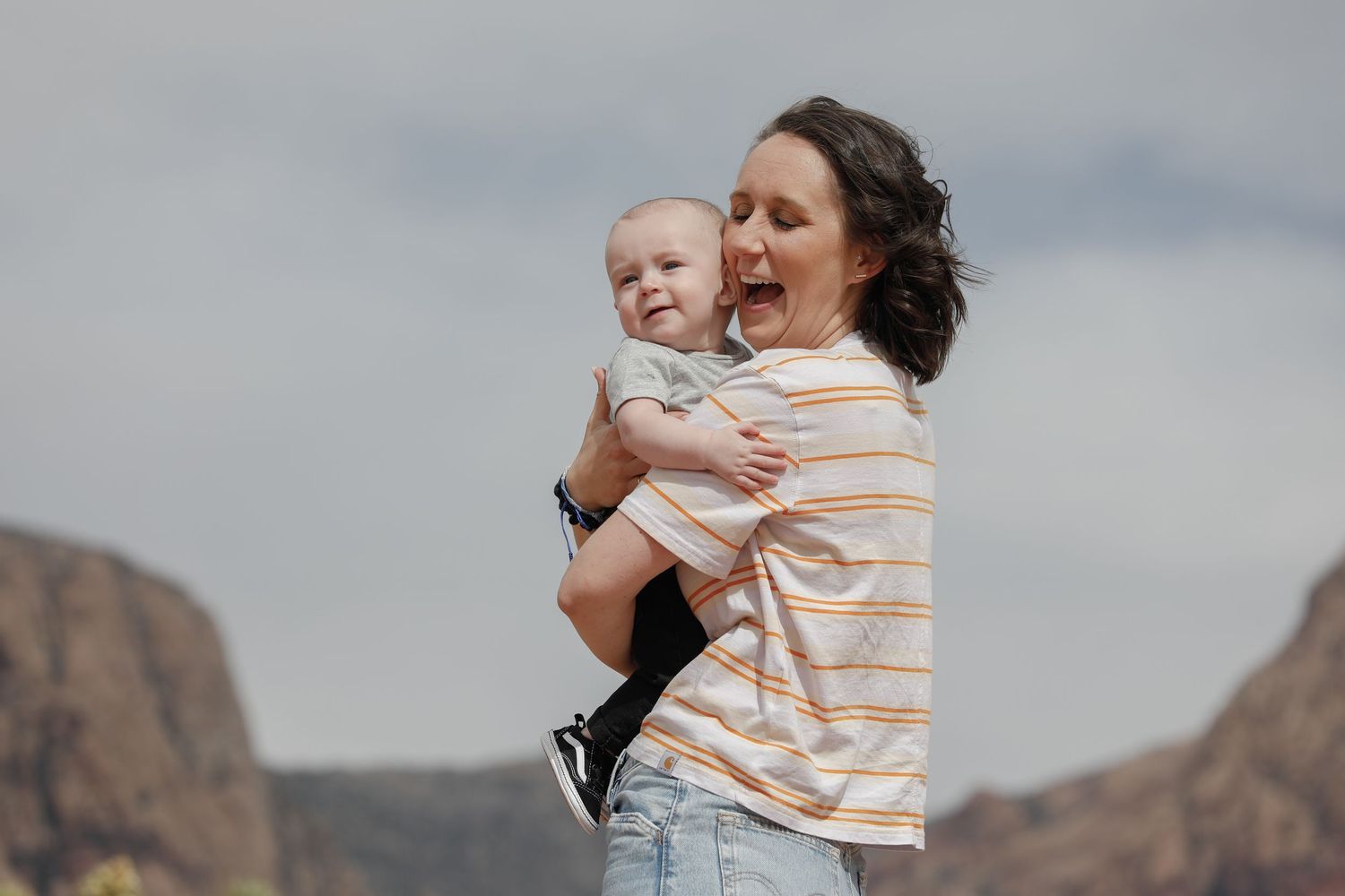 Parent and child share a joyful embrace against cloudy mountain backdrop.