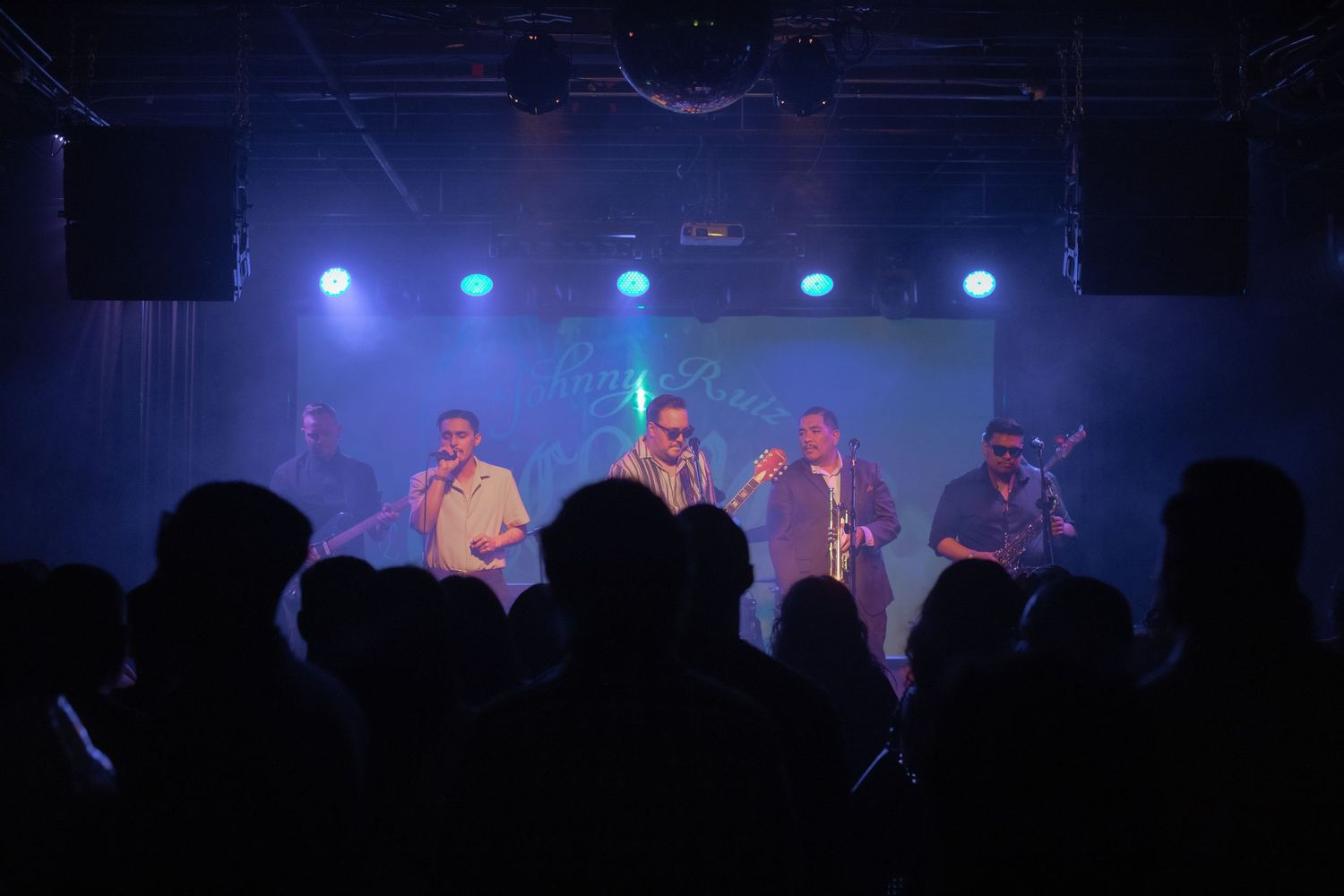 Band performs on stage with blue lighting and silhouetted audience in foreground.