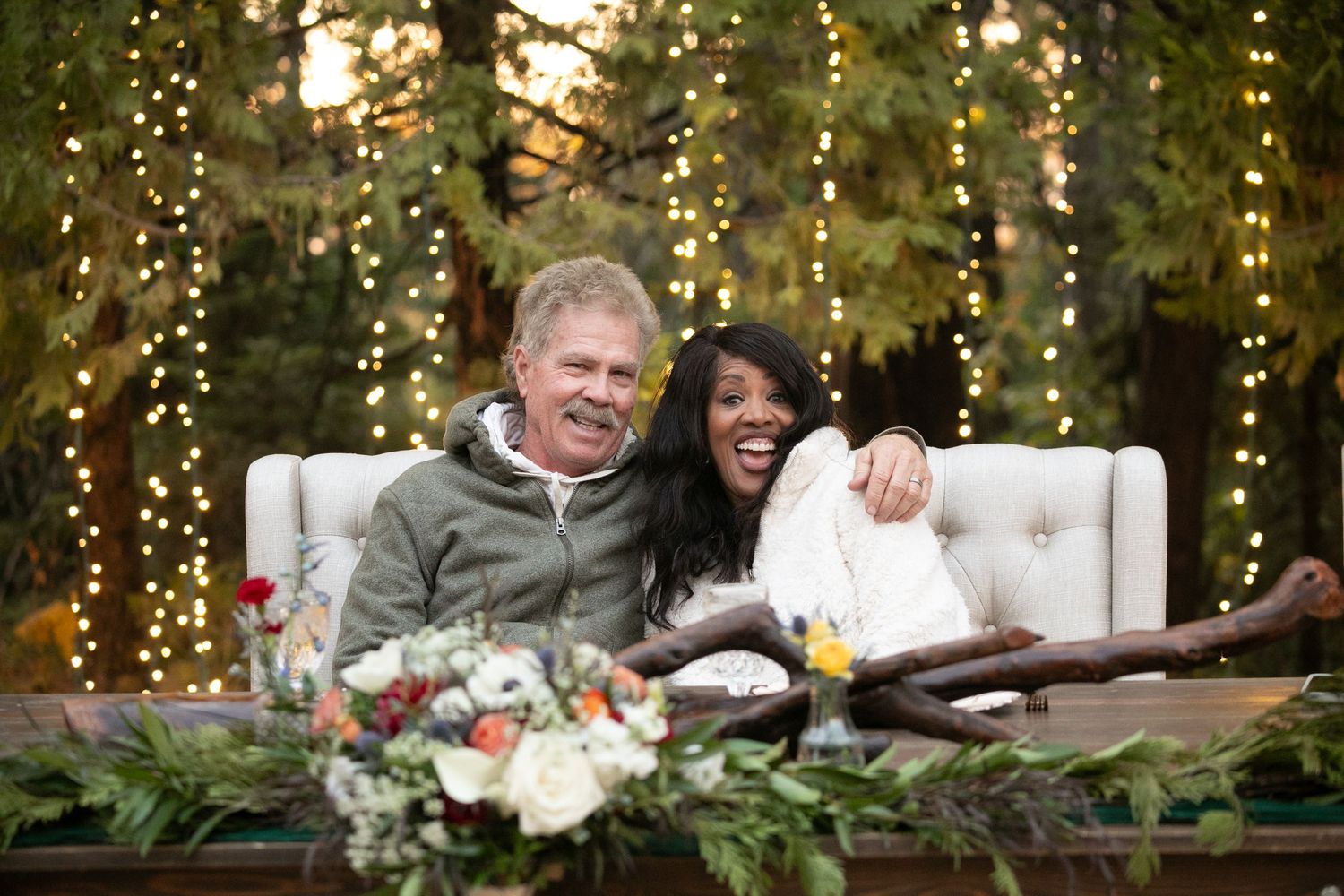 Wedding couple seated on white tufted couch with floral decorations and twinkle lights.
