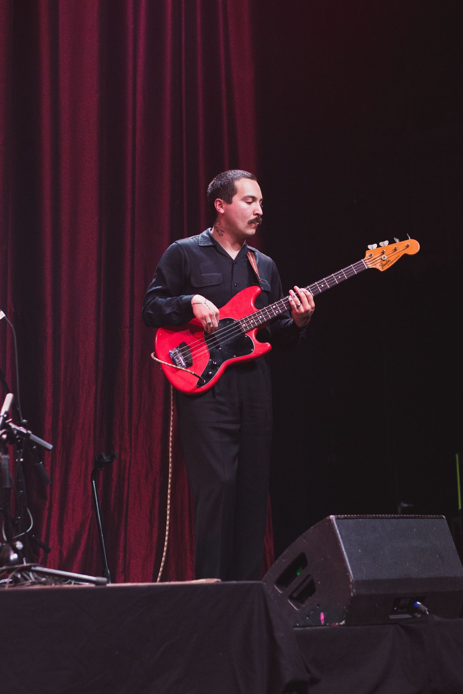 Bass guitarist in black attire performs on stage with a red bass guitar against burgundy curtains.
