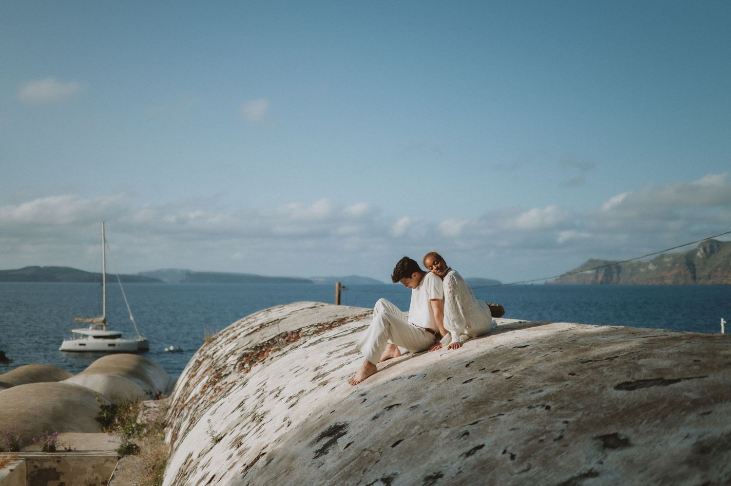 Couple sharing intimate moment on sun-bleached coastal rocks overlooking sailboat-dotted bay.