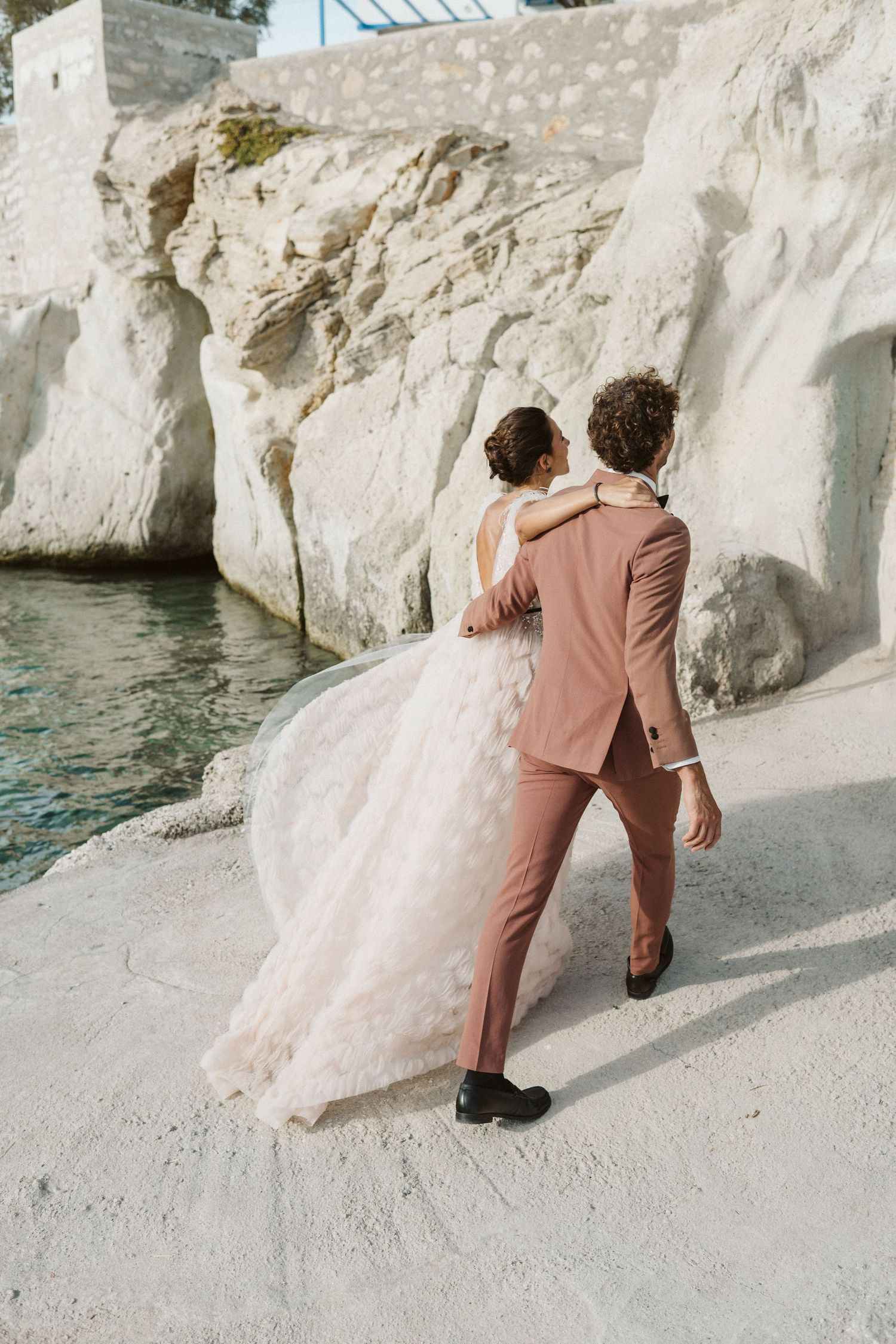 A couple in wedding attire stands together on white limestone rocks beside turquoise waters on a Mediterranean coastline.