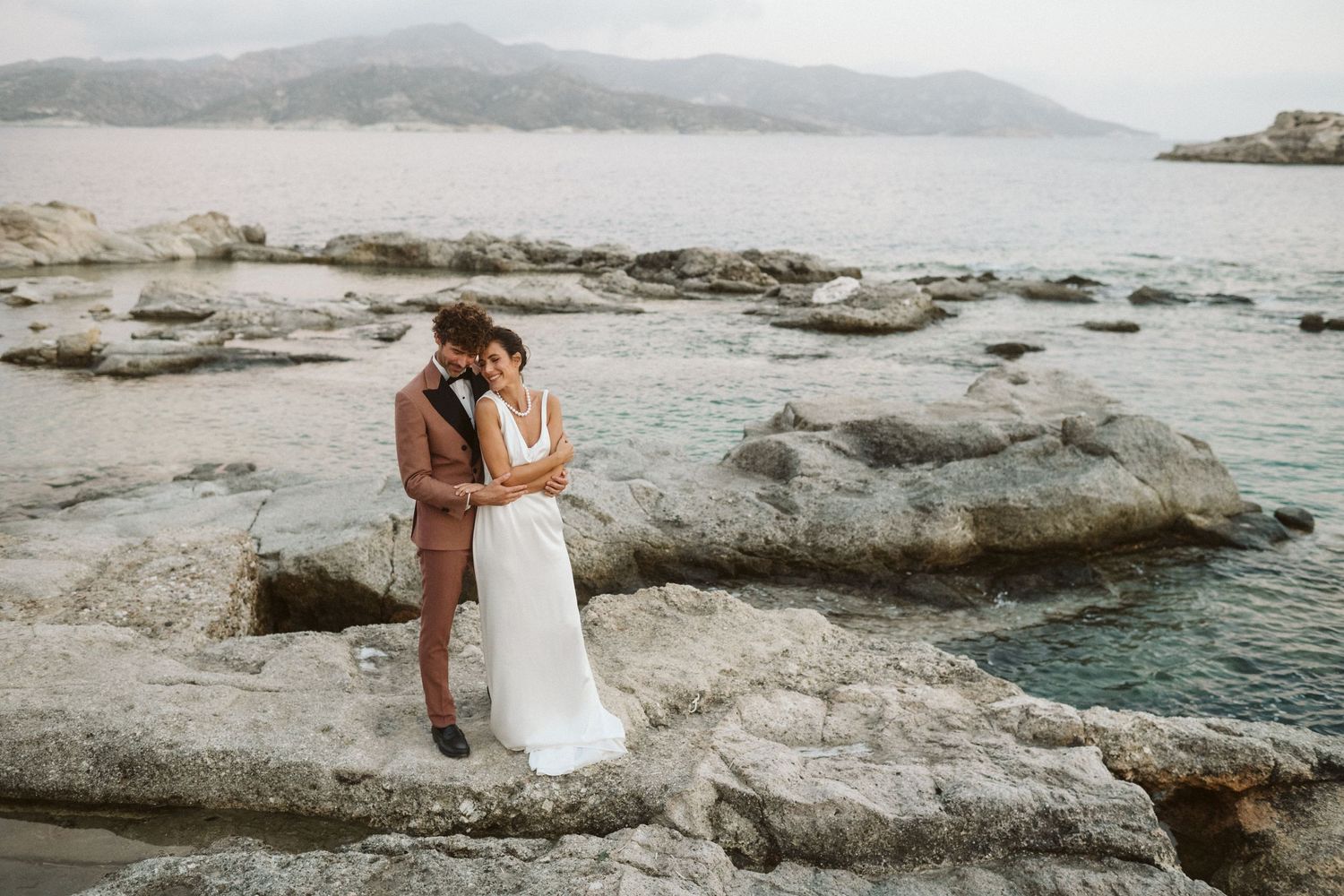 A couple embraces on rocky coastal outcropping overlooking misty ocean and mountains.