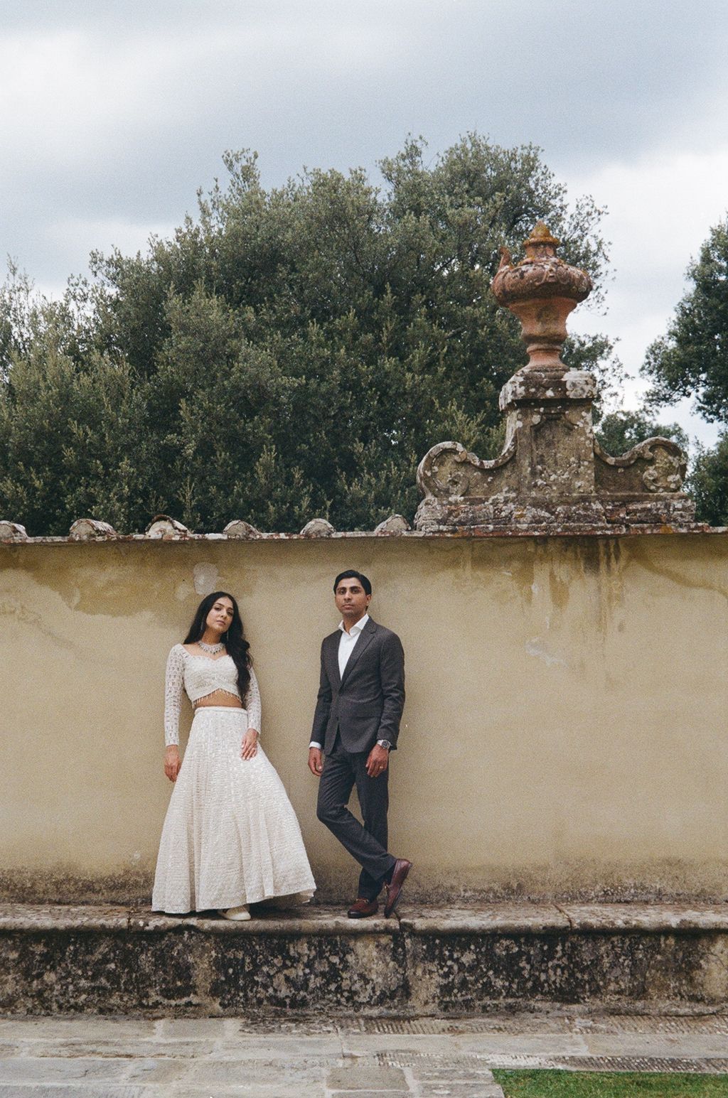 A couple in wedding attire poses against a beige stone wall with greenery in a romantic outdoor setting.