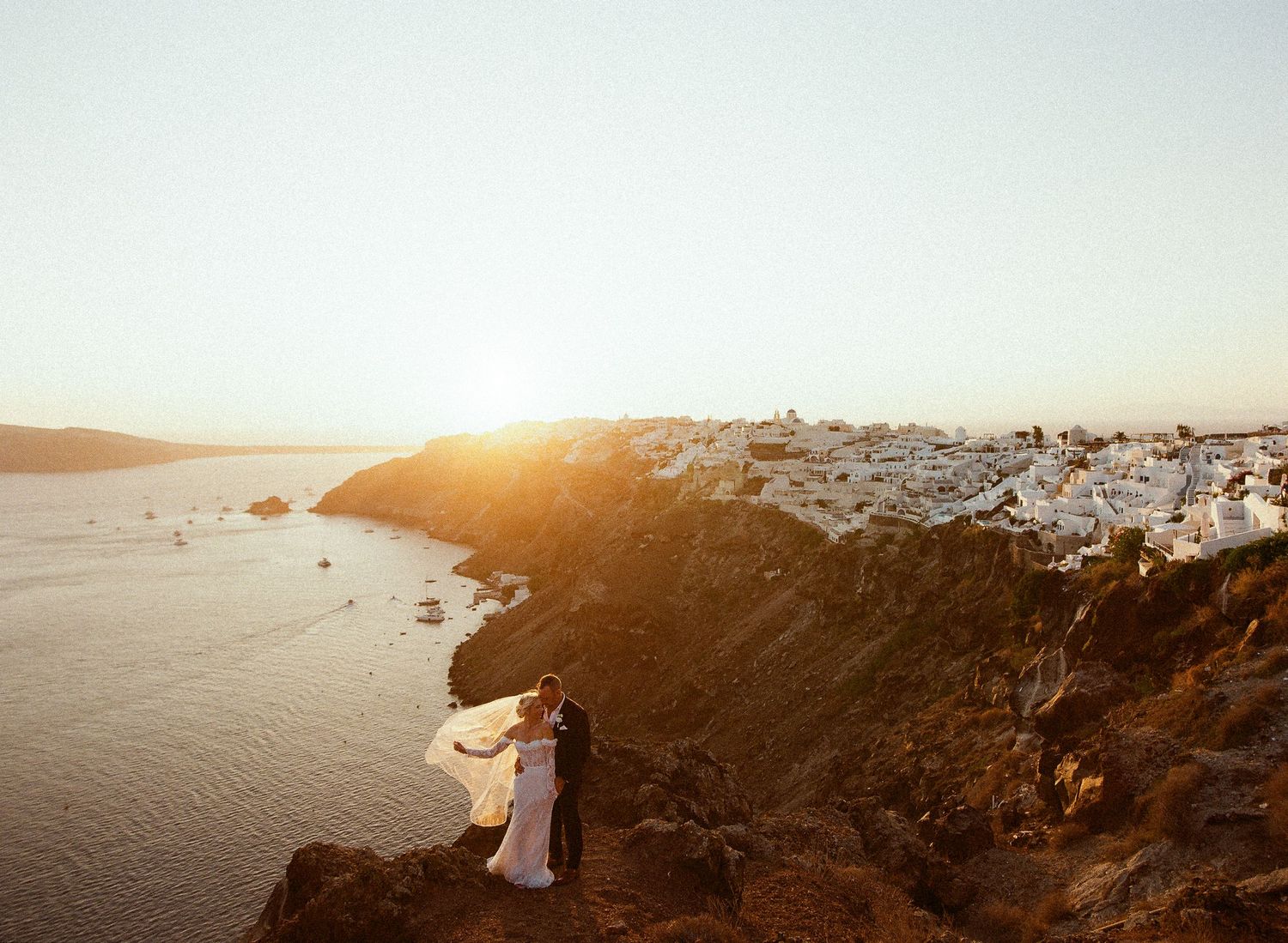 A couple stands on a cliff overlooking the white buildings of Santorini, Greece during golden sunset over the Aegean Sea.