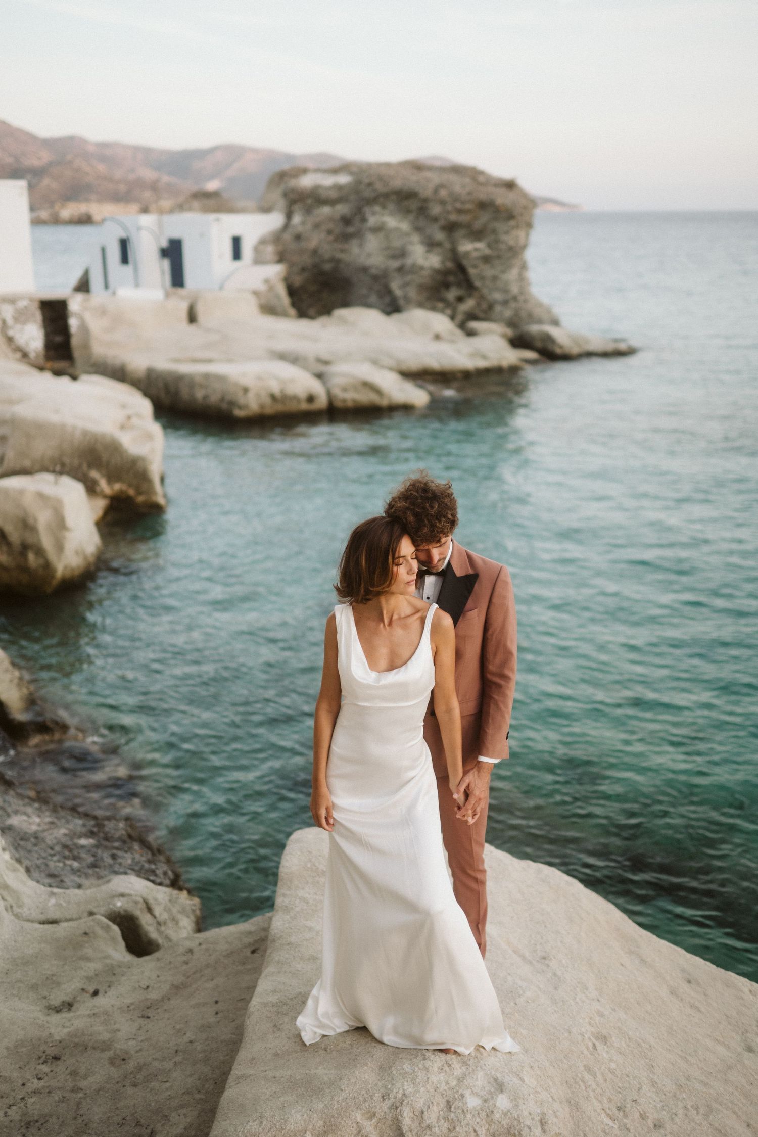 A couple stands on coastal rocks overlooking turquoise Mediterranean waters near white cliffside buildings.