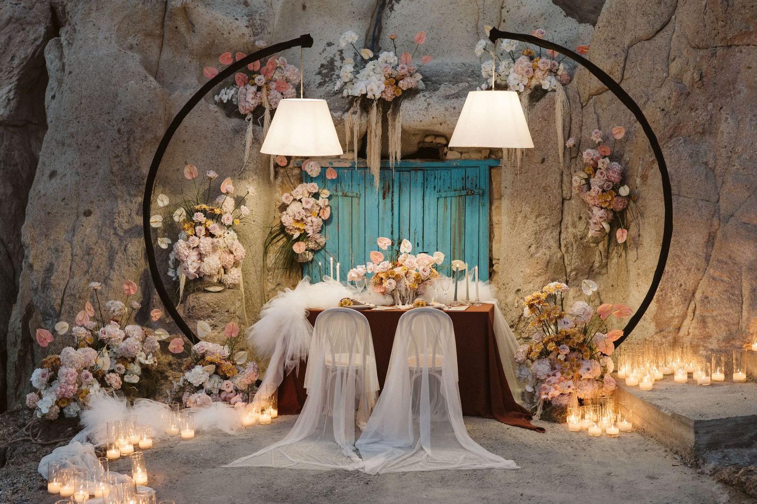 Romantic wedding ceremony setup with round arch, white chairs, teal curtains, and floral decorations illuminated by candlelight.