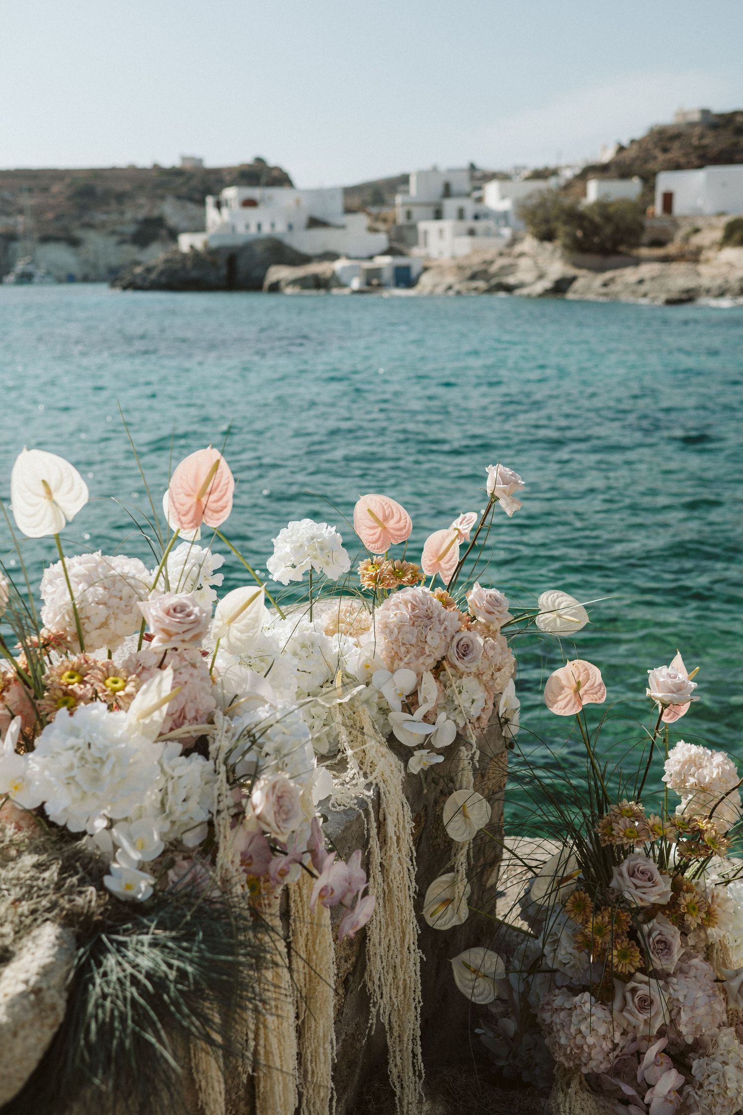 Pink and white flowers arranged on a rock overlooking turquoise Mediterranean waters with white coastal buildings in Greece.