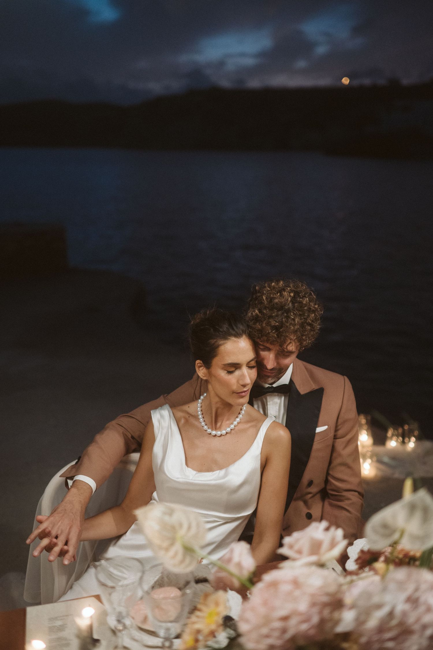 Couple shares an intimate candlelit dinner moment by the lake at night with pink floral table decorations.
