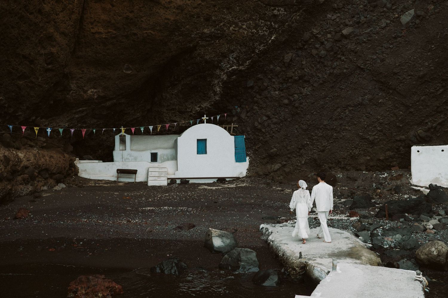 Two figures in white walk near a small white church building against dark rocky cliffs.