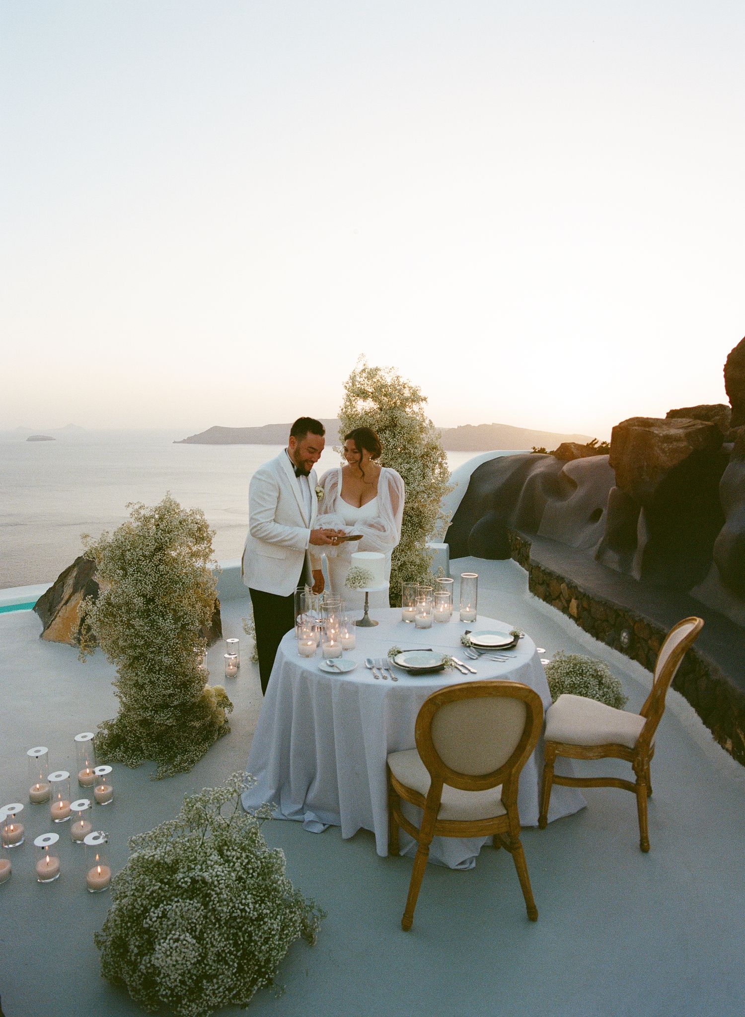 A romantic dinner setup with white floral arrangements on a seaside terrace in Santorini at sunset.