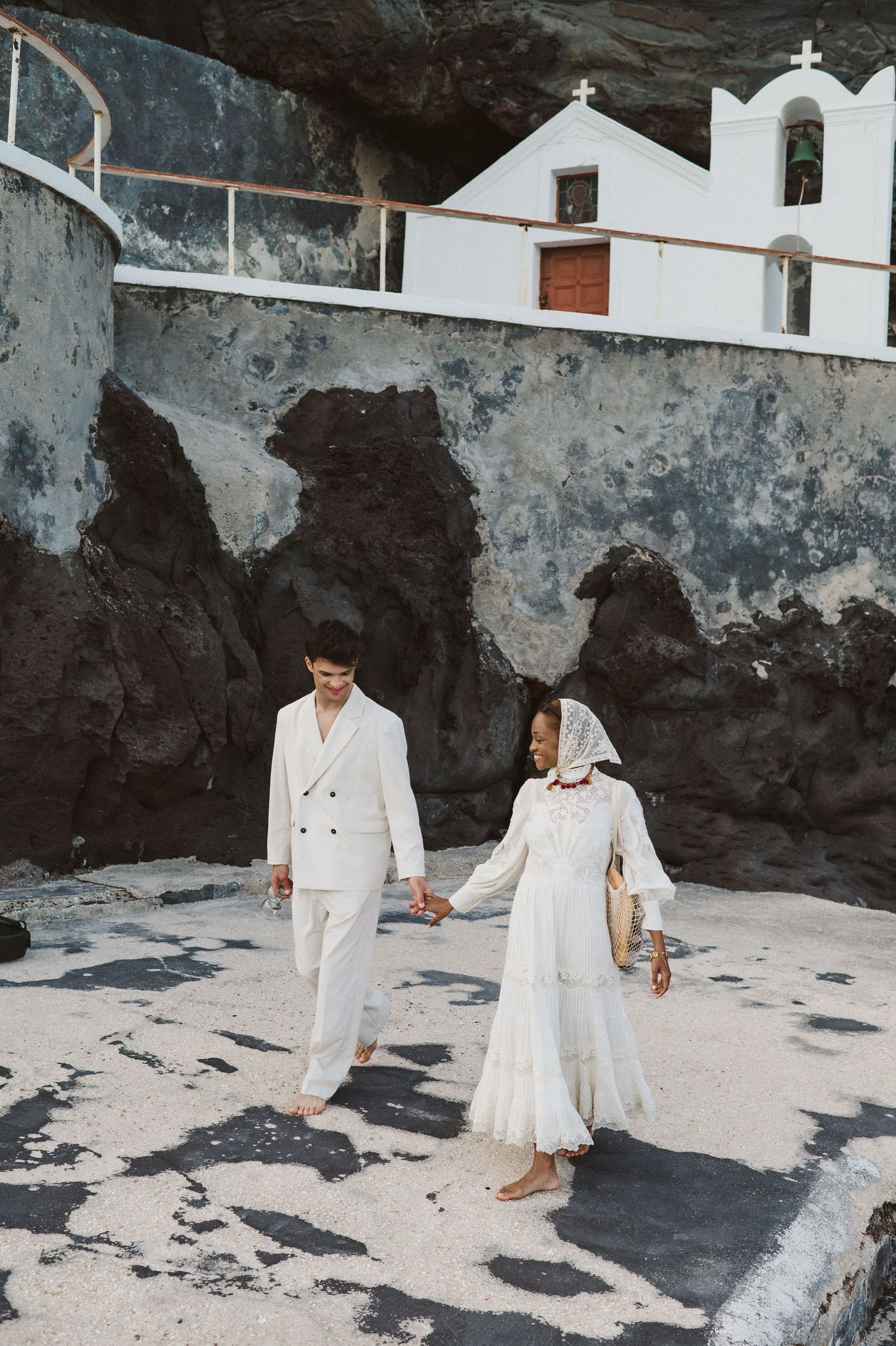 A couple walks hand in hand near a whitewashed church against dramatic rocky cliffs.