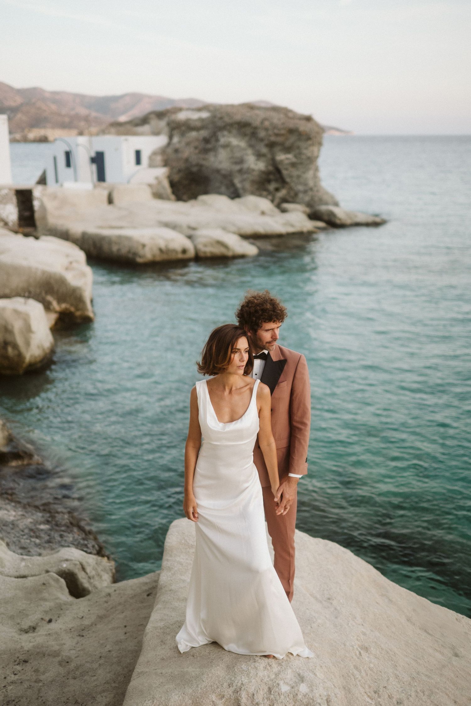 A couple stands on white coastal rocks overlooking turquoise Mediterranean waters near traditional Greek island architecture.