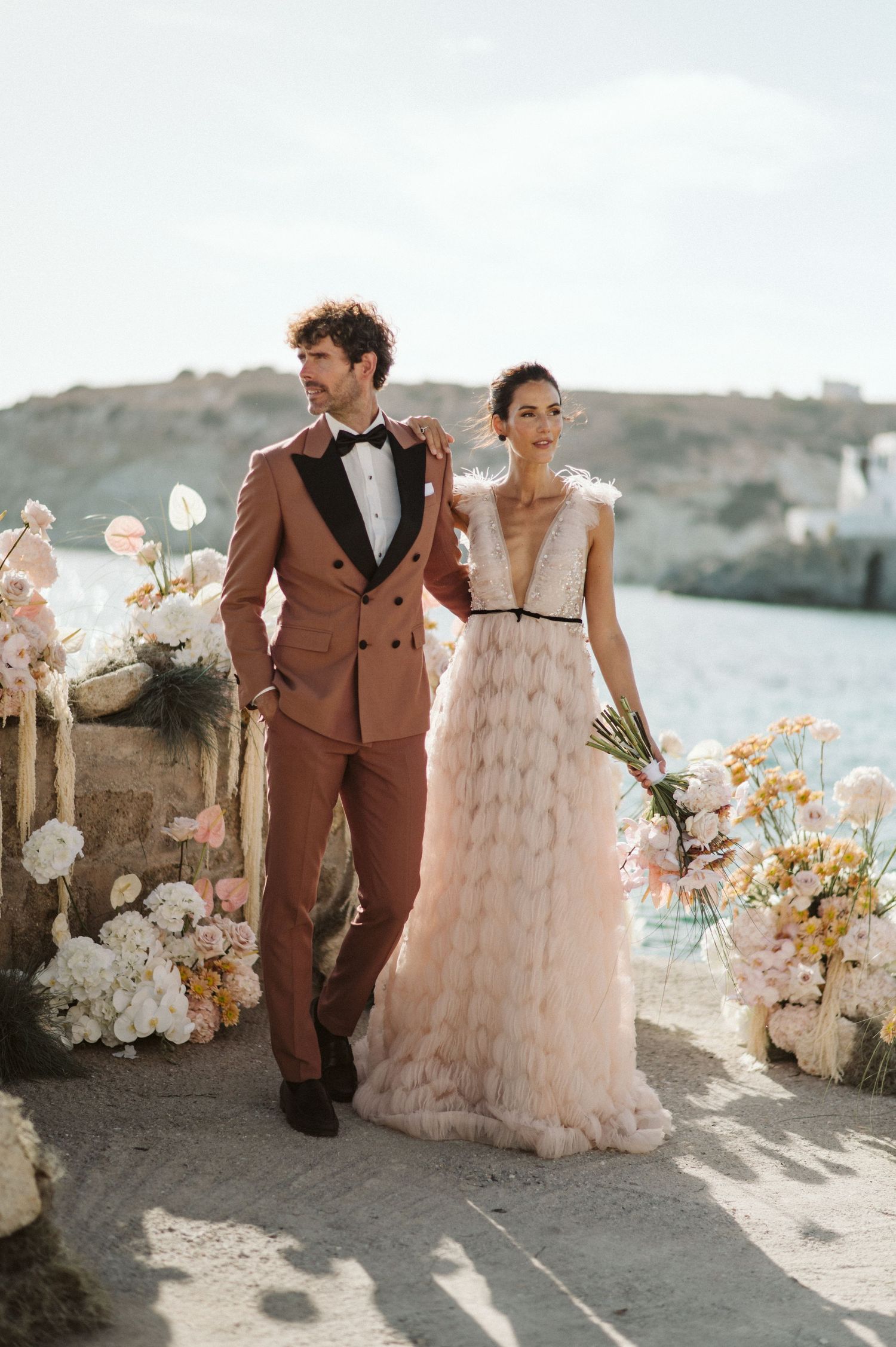Elegant couple in wedding attire, with brown suit and blush tulle gown, pose by floral arrangements near seaside.
