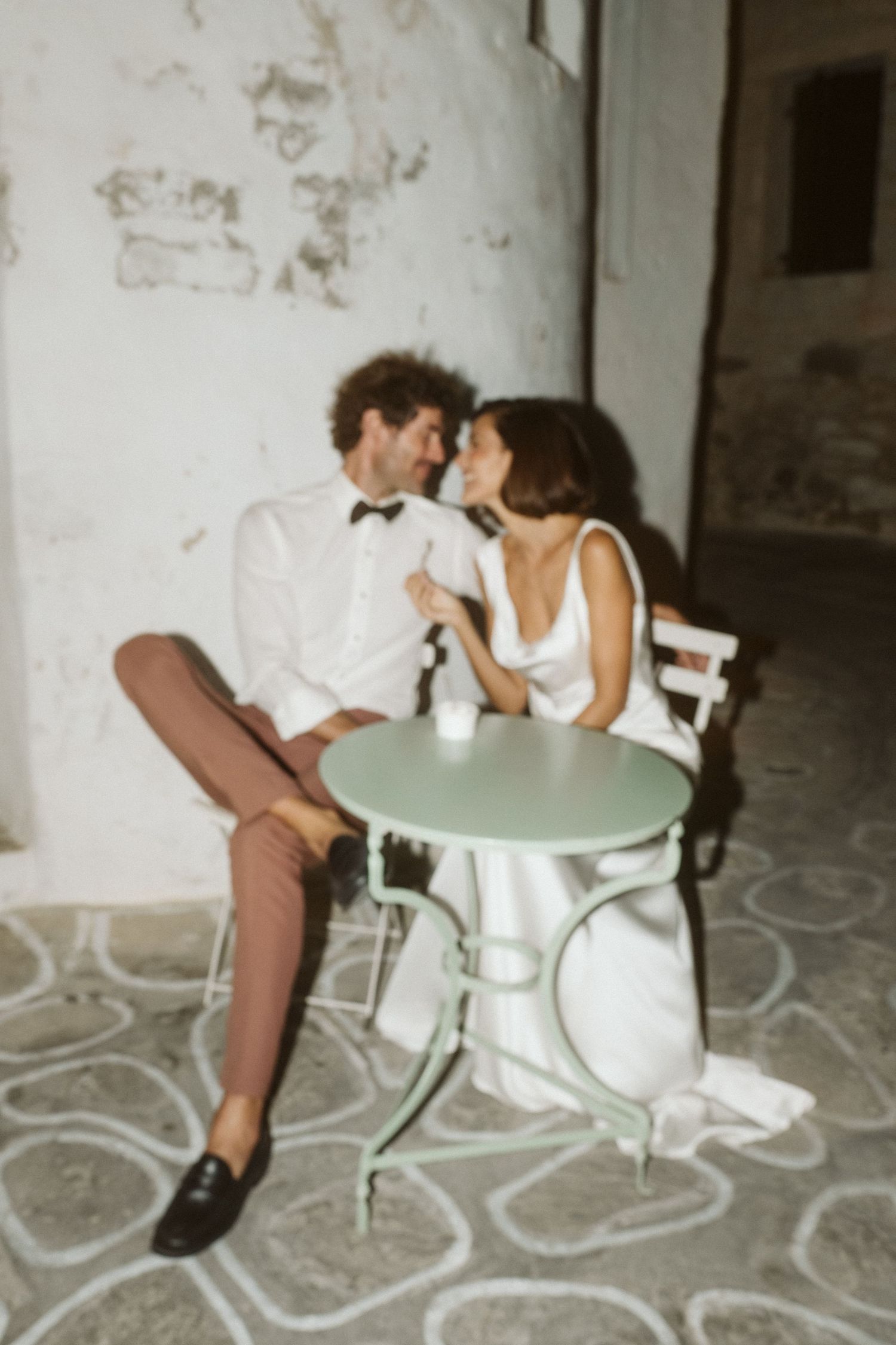 A couple in wedding attire shares an intimate moment at a small mint green cafe table while sitting outdoors.
