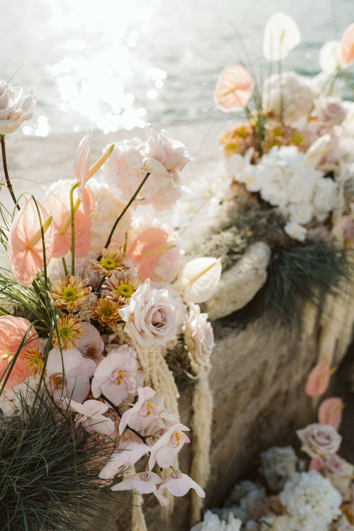 Romantic floral arrangement featuring soft pink roses, orchids and wildflowers in natural sunlight against a textured backdrop.