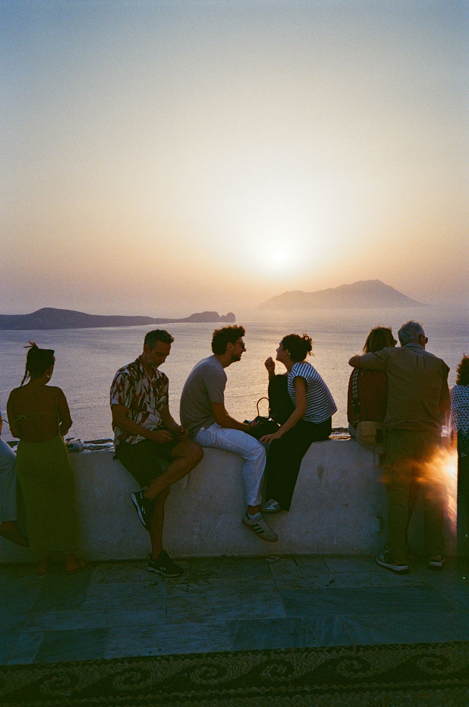 Silhouettes of people sitting on a wall at sunset overlooking a coastal mountain landscape.