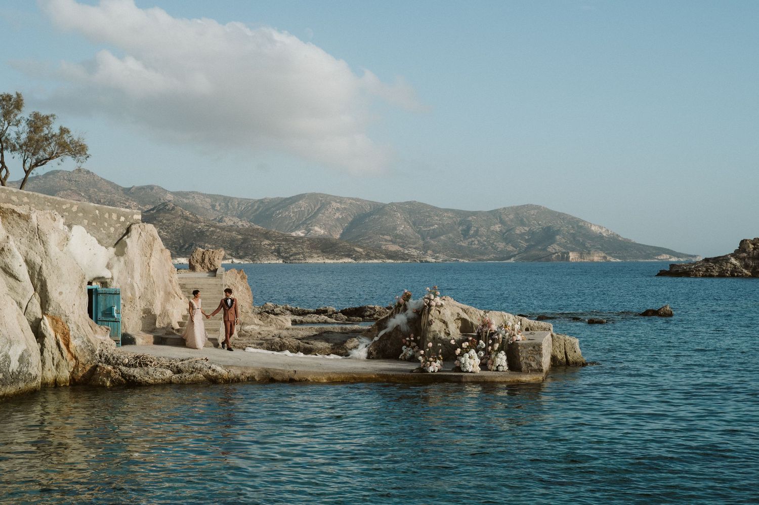 Small white Mediterranean coastal structure built into rocky shore with blue sea and mountains in background.