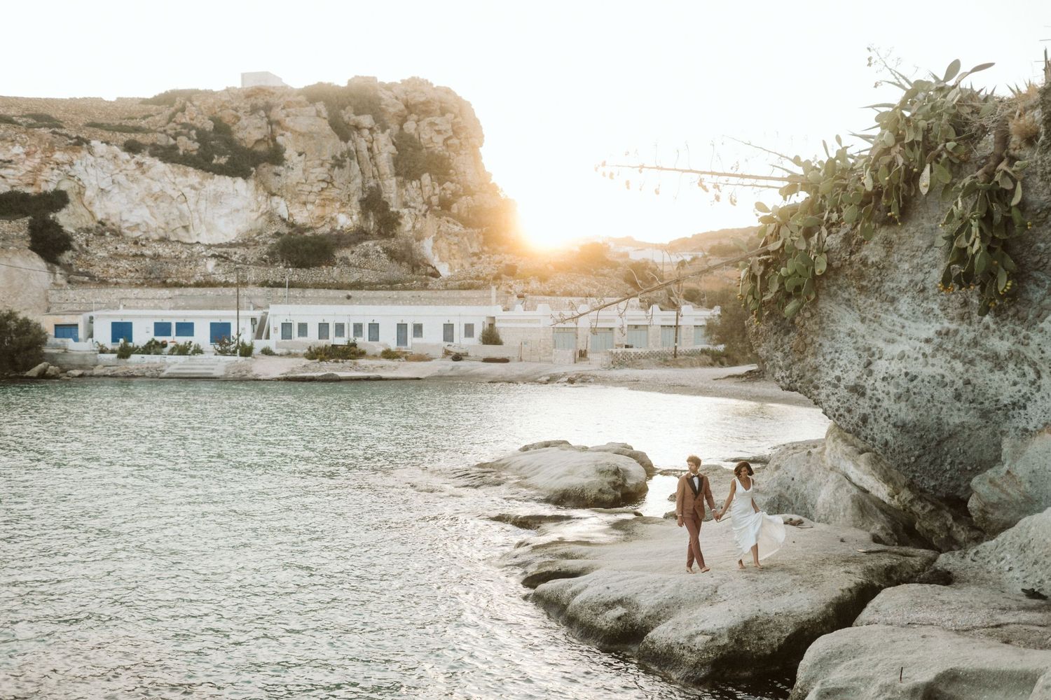 A silhouette stands on a white rocky cliff overlooking a Mediterranean beach cove at sunset with coastal buildings below.