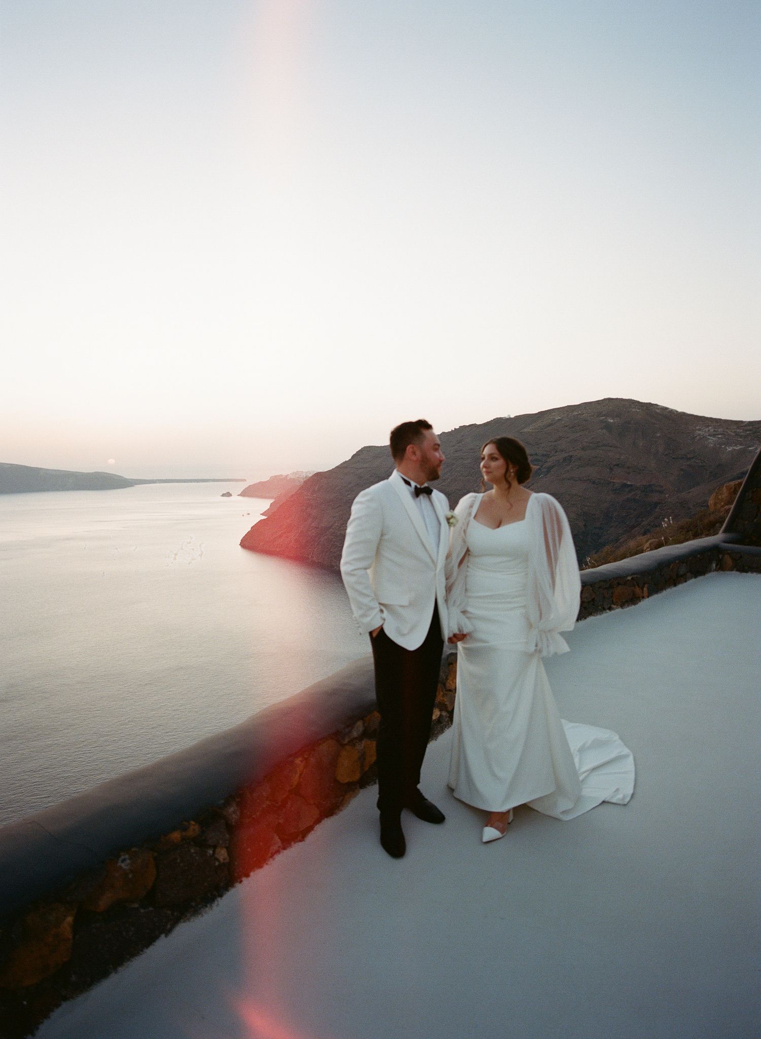A couple in wedding attire stands on a white terrace overlooking the Mediterranean Sea at sunset in Santorini, Greece.