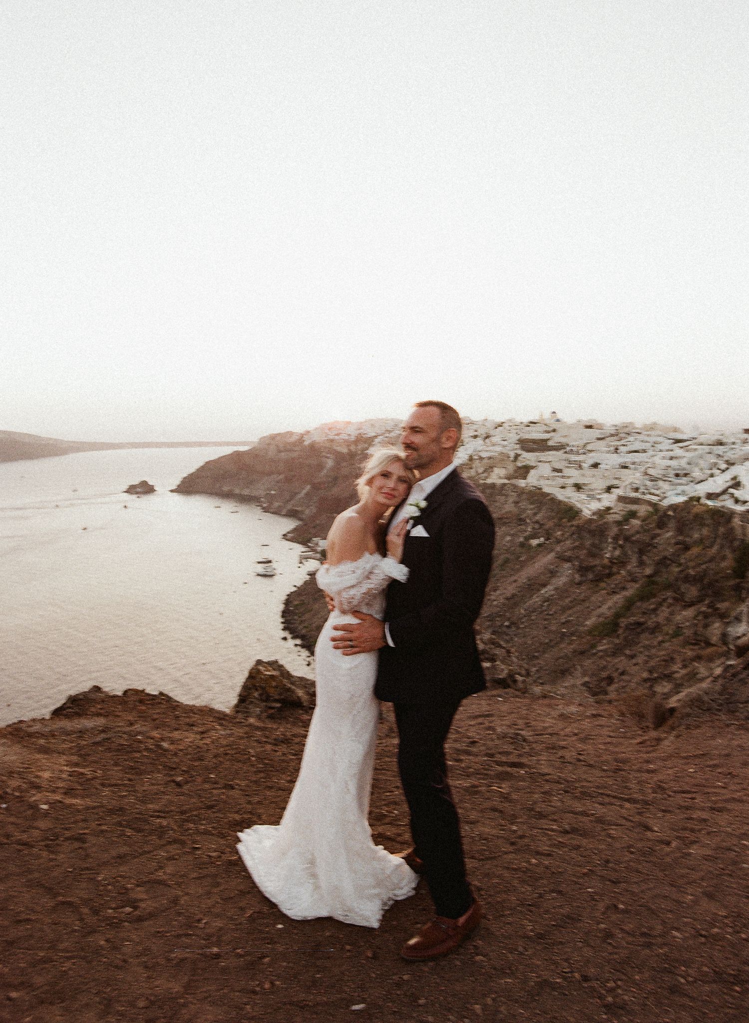 A couple in wedding attire embrace on a cliff overlooking a scenic coastal view at sunset.