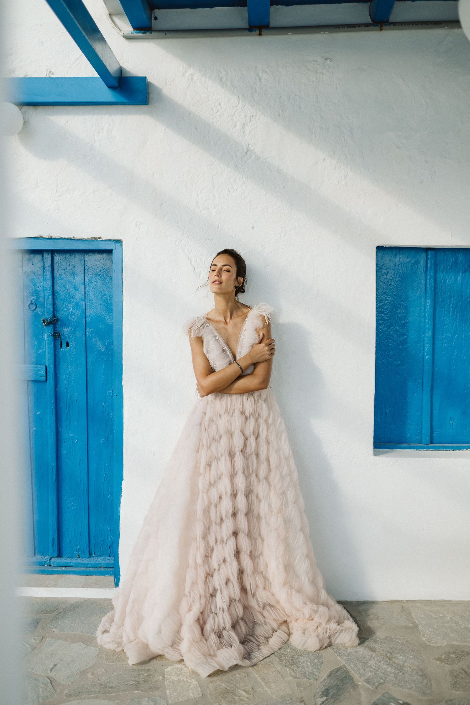 A figure in a flowing blush tulle gown poses against a white wall with bright blue shutters and beams.
