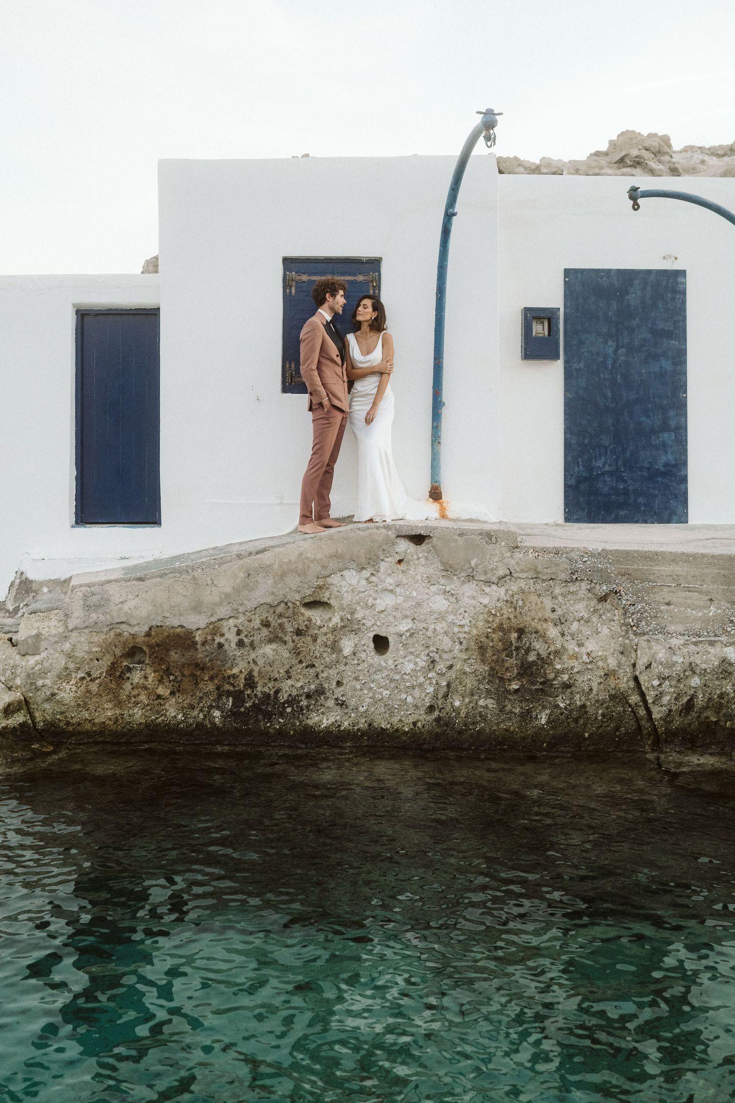 A couple in wedding attire stands embracing against a white Mediterranean building with blue doors beside turquoise waters.