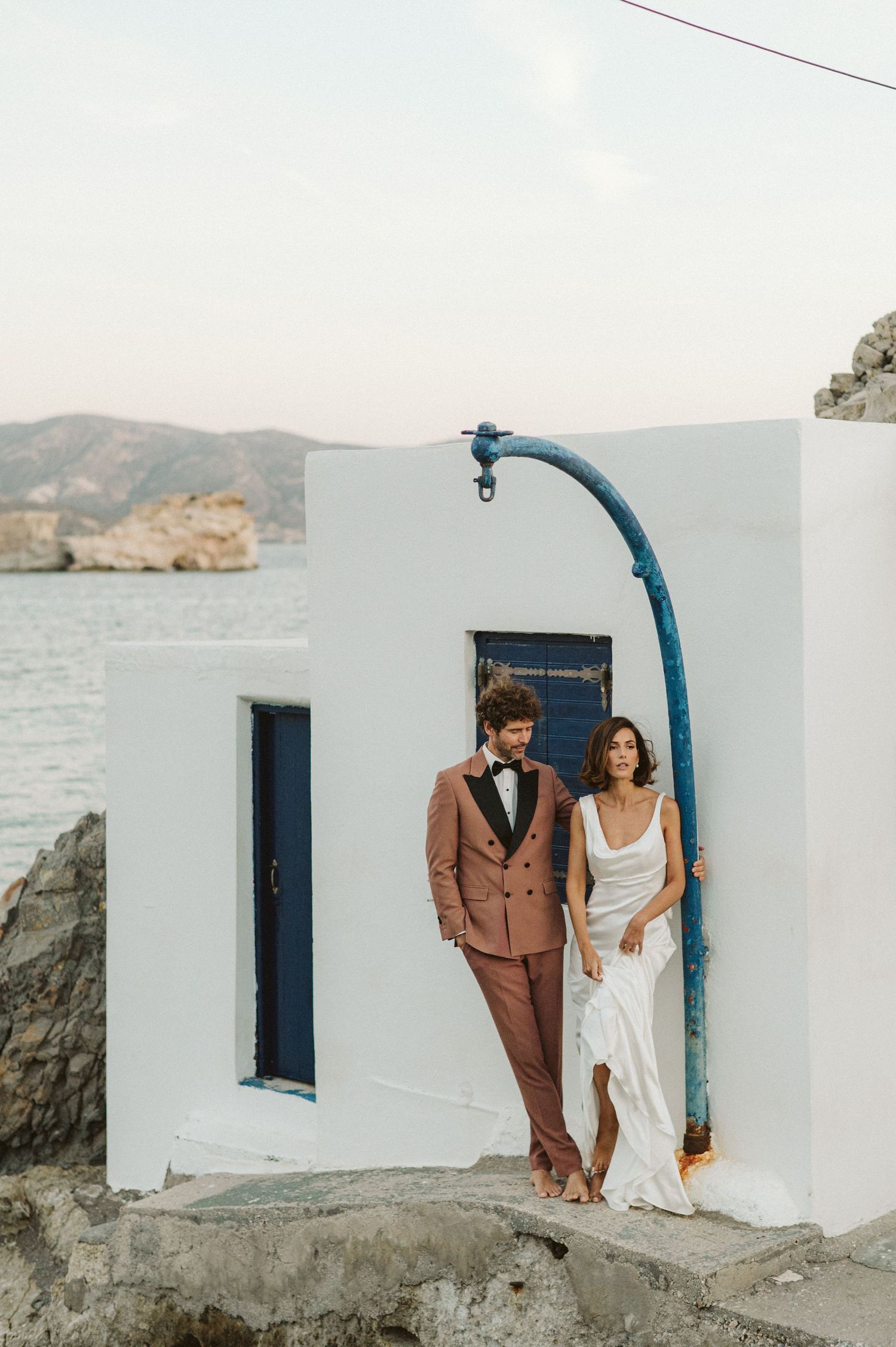 A couple in wedding attire stands against a white Mediterranean building with blue accents overlooking the ocean.
