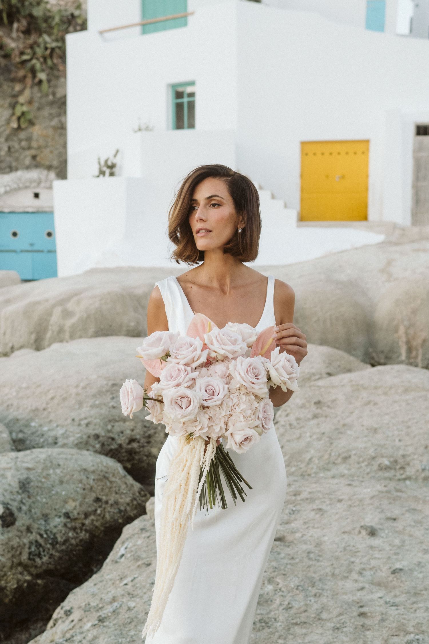 Elegant bride in white dress holds beautiful bouquet of pink roses against white Mediterranean building backdrop.