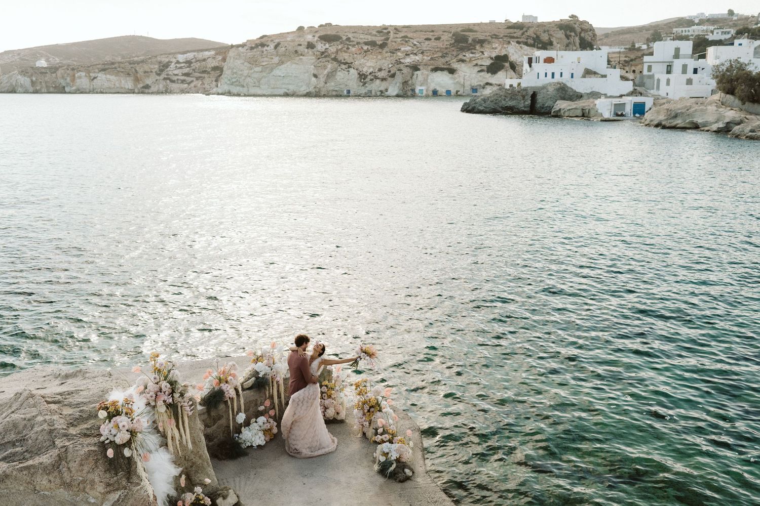 A couple sits on rocky coastal outcrop overlooking Mediterranean waters with white buildings dotting the shoreline.