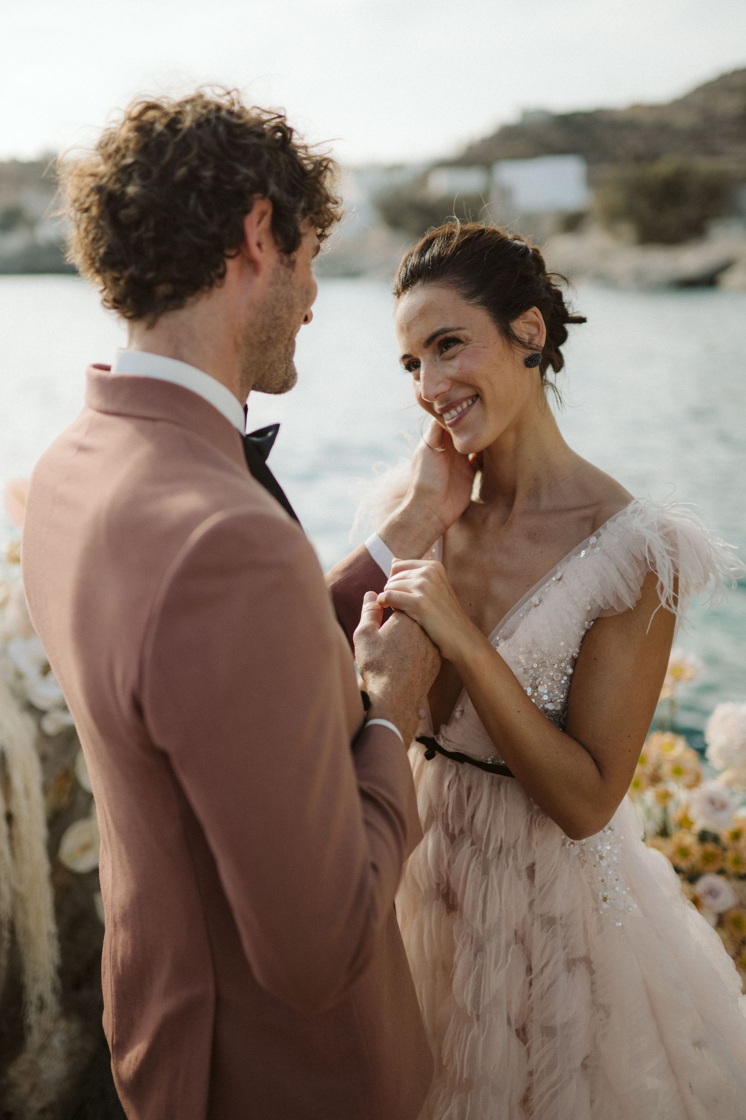 A couple shares a tender moment in formal wedding attire against a coastal backdrop during golden hour.