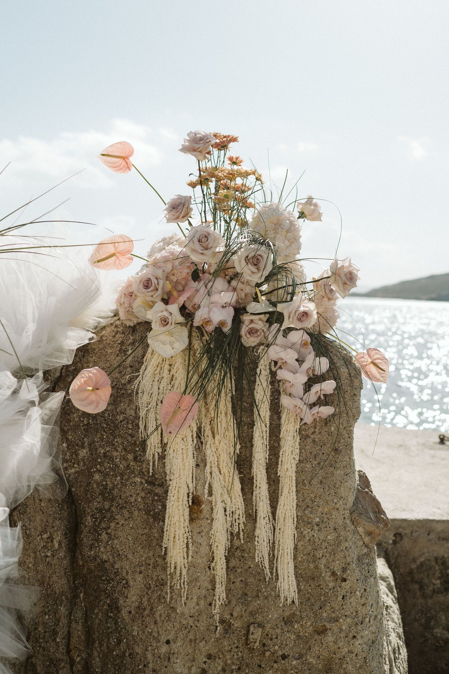 Elegant floral arrangement with dried flowers and pampas grass displayed on a rock by the ocean with soft peach and neutral tones.