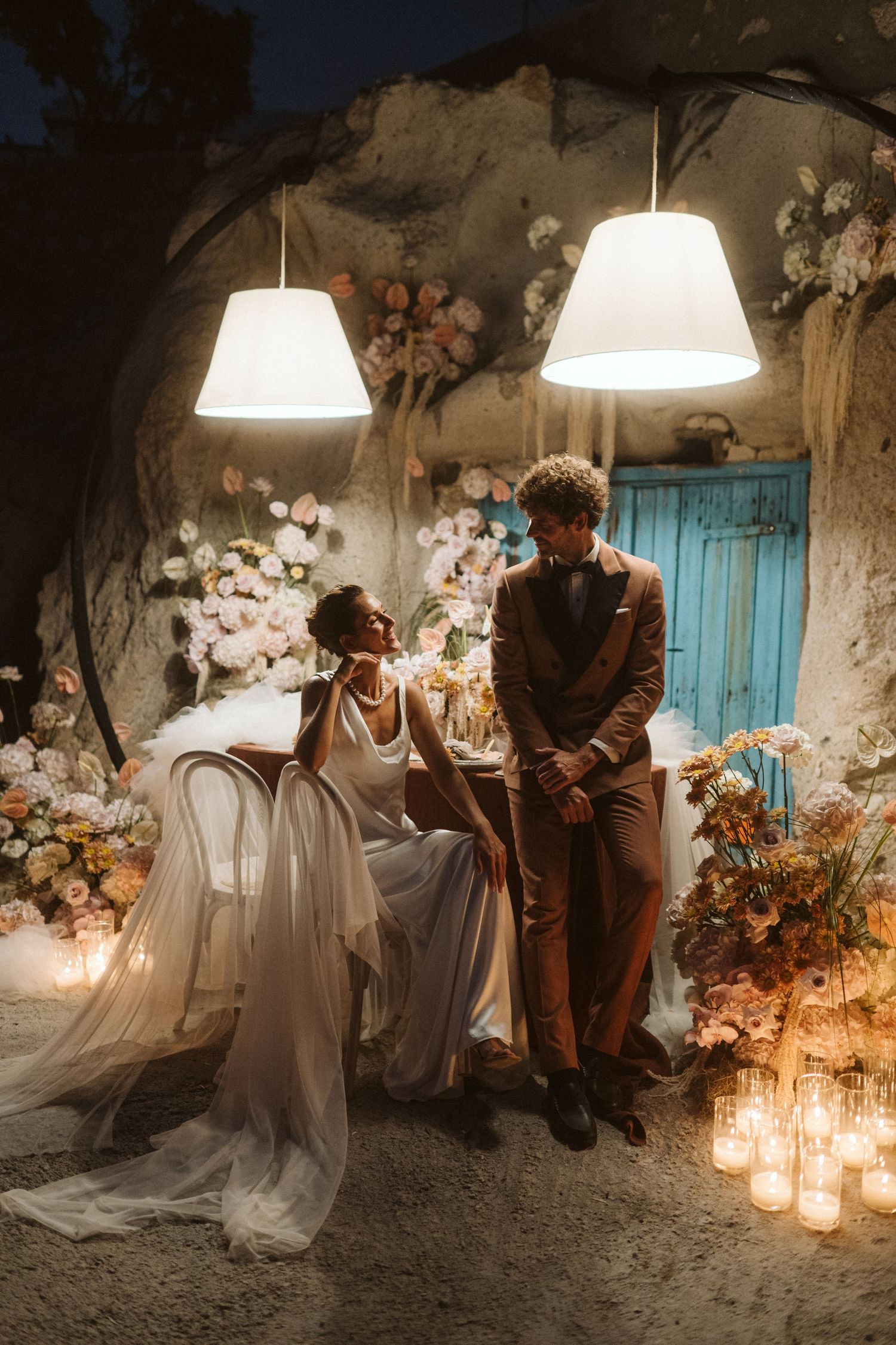 A romantic wedding scene with two people seated among candles and floral decor under pendant lights with a blue door backdrop.