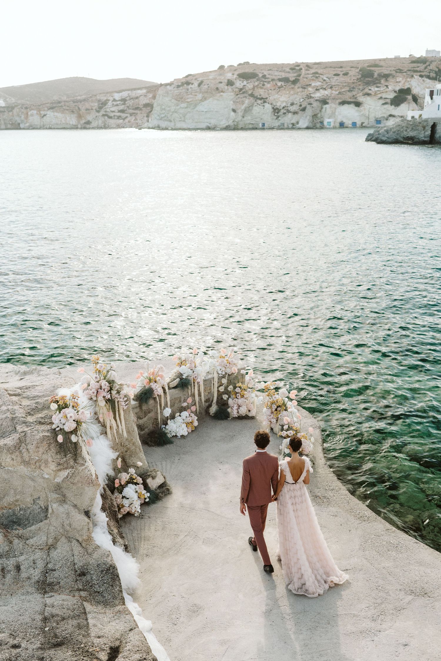 Couple standing near floral wedding arch overlooking turquoise Mediterranean waters and white cliffs on seaside cliff.