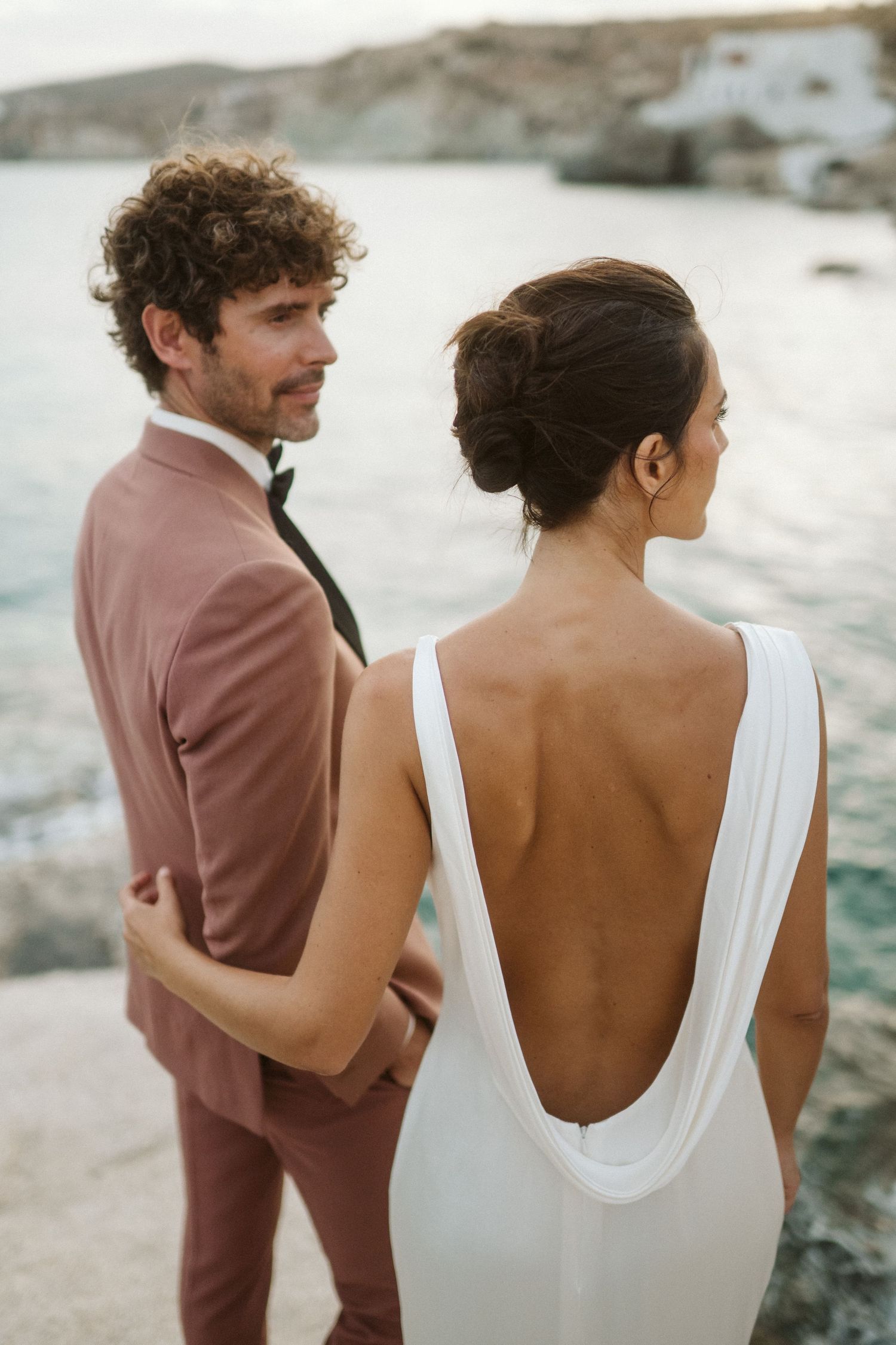 Couple in romantic wedding attire pose by rocky coastal shore, bride in backless white dress and groom in dusty rose suit.